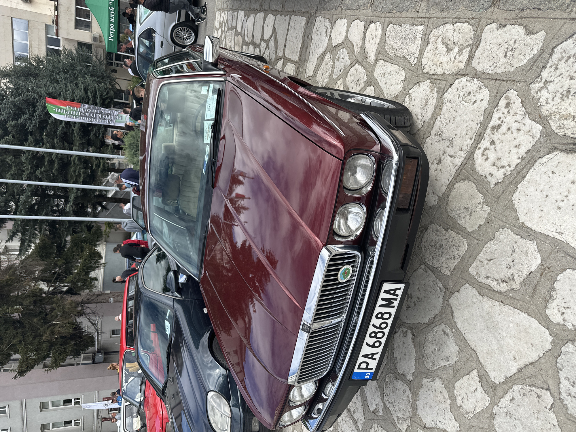 A front-facing shot of a maroon Jaguar with the Bulgarian license plate "PA6868MA" on a stone-paved street. The location is likely Razlog, Bulgaria, given the Bulgarian license plate. Other cars, in black and red, are partially visible behind the Jaguar. People are gathered in the background near a building, with what appears to be a banner or flag present. The environment suggests an outdoor gathering or car meet in an urban setting.