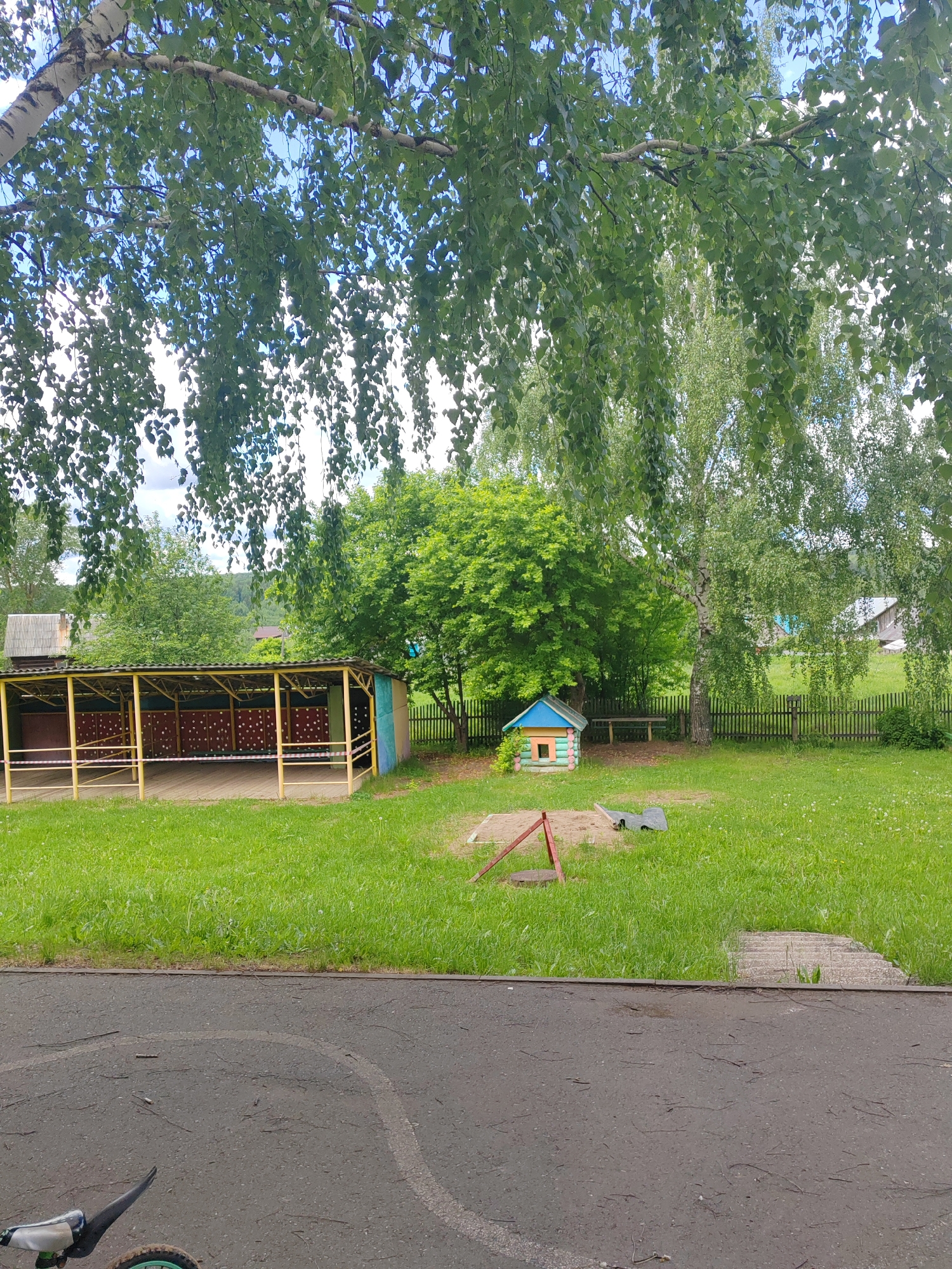 The image captures a playground or recreational area in Unknown, Unknown, with features characteristic of a park or community space. A partially visible paved area in the foreground gives way to a grassy space containing a small, colorful playhouse, a sandbox, and other playground equipment. A covered structure with a wooden framework is visible on the left side of the image, possibly for shelter or seating. In the background, a wooden fence separates the playground from a more wooded area with various trees. The upper portion of the image shows the canopy of trees, suggesting a natural and somewhat secluded setting. Overall, the scene is a representation of a public space intended for recreation and leisure.