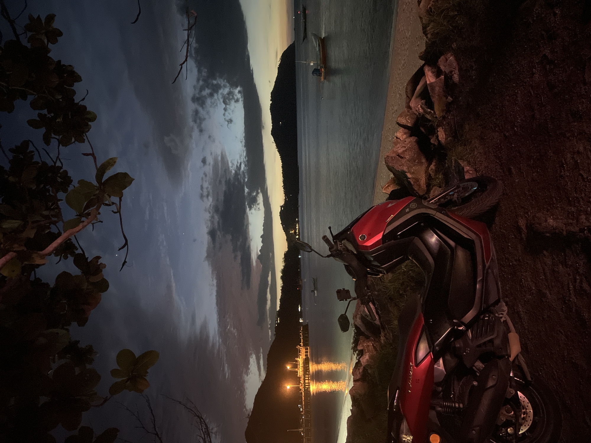A red scooter is parked on a dirt and gravel area next to a rocky beach. The scooter is in the foreground, positioned diagonally from the lower left to the middle right of the frame. Behind the scooter, the sea stretches out to a distant shoreline with hills. The sky is dark blue with scattered clouds, indicating it is either dusk or dawn. Lights from a pier and buildings can be seen on the left side of the scene, reflecting on the calm water. Several small boats are present in the water, some further out in the bay. Branches of a tree with dark leaves hang over the top of the image, obscuring parts of the sky. The overall scene depicts a peaceful coastal setting at twilight.
