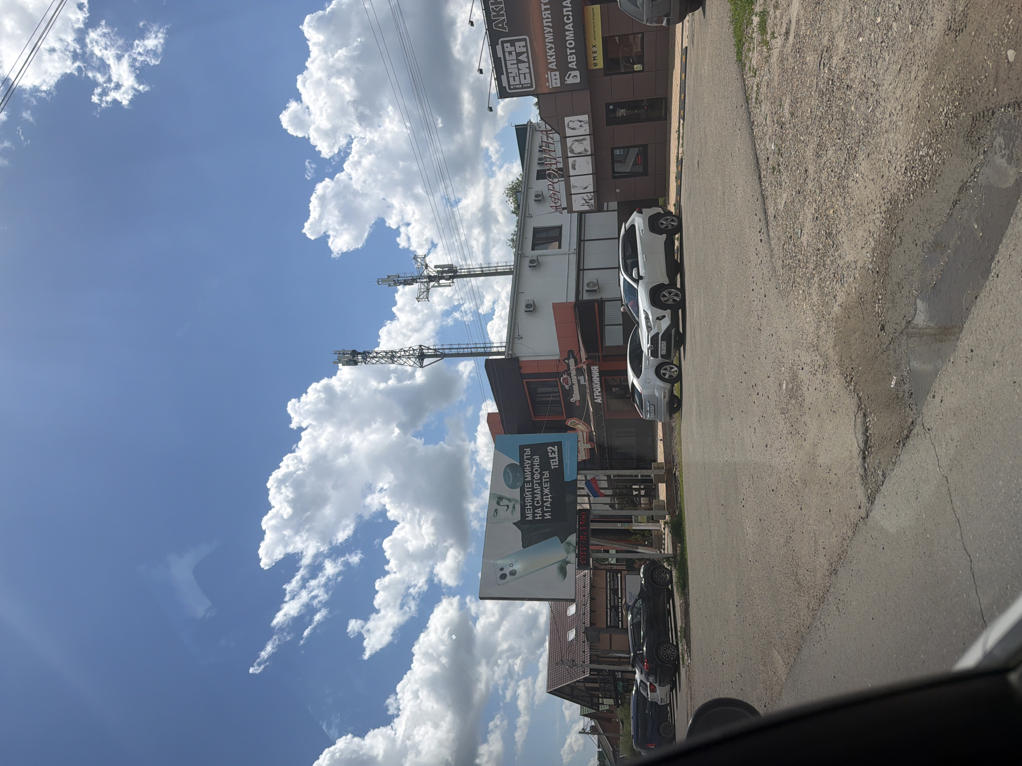 This is a street-level, eye-level, medium shot of a commercial strip in Leningradskaya, Russia, on a sunny day with scattered clouds. The street is paved with asphalt and shows signs of wear, including potholes and gravel. Several cars are parked along the roadside. On the right side of the image, a building with multiple storefronts is visible. One storefront has a sign that reads "СУПЕР СИЛА" (Super Strength) above a sign that advertises "АККУМУЛЯТОРЫ" (Batteries) and "АВТОМАСЛА" (Motor Oils). Another sign reads "EMEX". Further to the left, a building with a sign reading "АФРОДИТА" (Aphrodite) is visible, with images of women's faces above the entrance. Another building displays a large billboard advertising "TELE2" and text in Russian, which translates to "Exchange minutes for smartphones and gadgets." Behind these buildings, tall cell towers rise against the sky. The left side of the image shows houses with pitched roofs. The overall impression is a typical suburban or small-town commercial area.