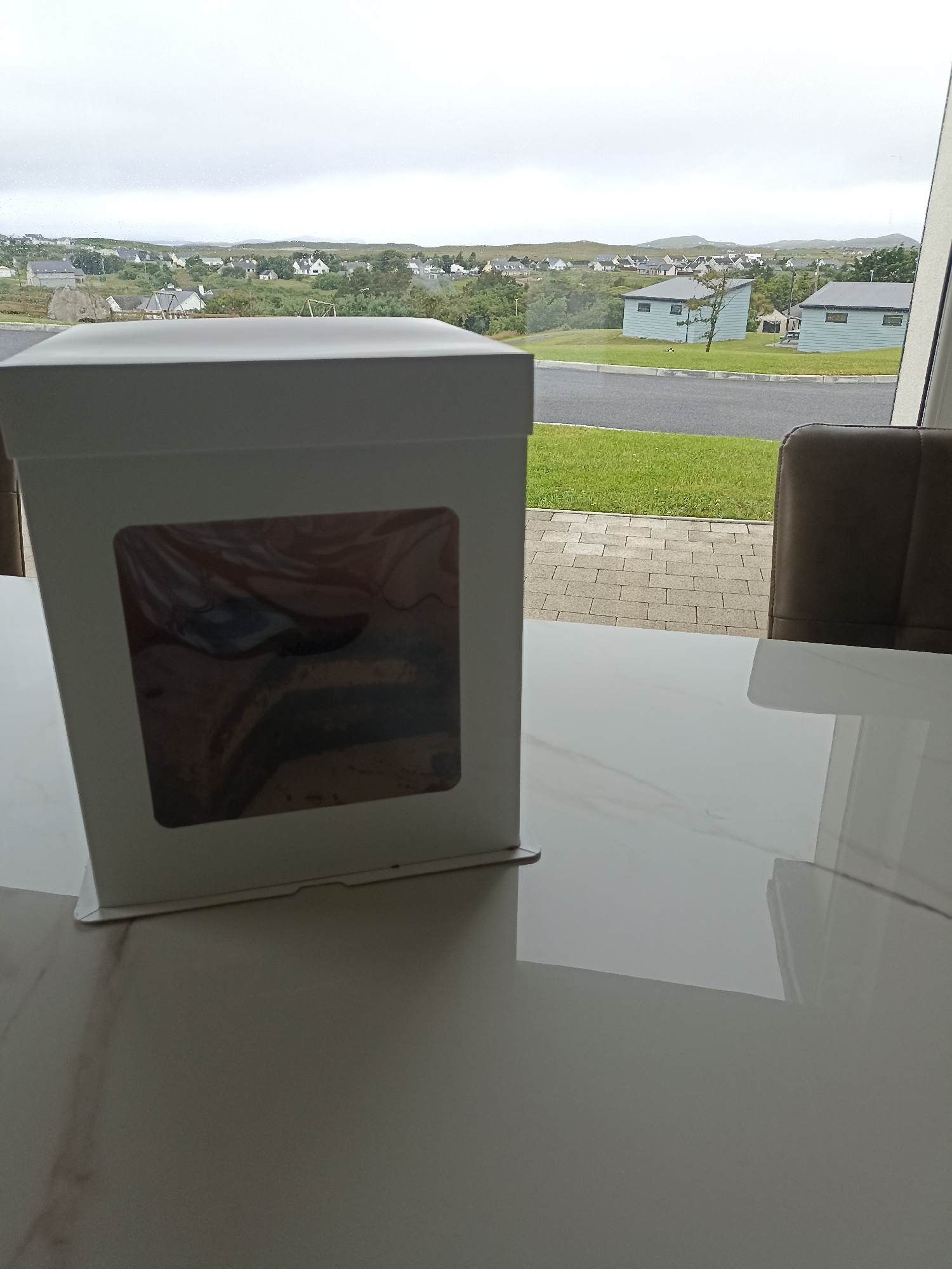 A white cake box with a window is placed on a white marble table. The box contains a layered cake, partially visible through the window. In the background, through a window, a scenic view of a village in Doirí Beaga, Ireland, is visible, with houses scattered across rolling green hills under an overcast sky. A brown leather chair is partially visible to the right of the cake box. The overall scene suggests an indoor setting looking out onto a picturesque landscape.