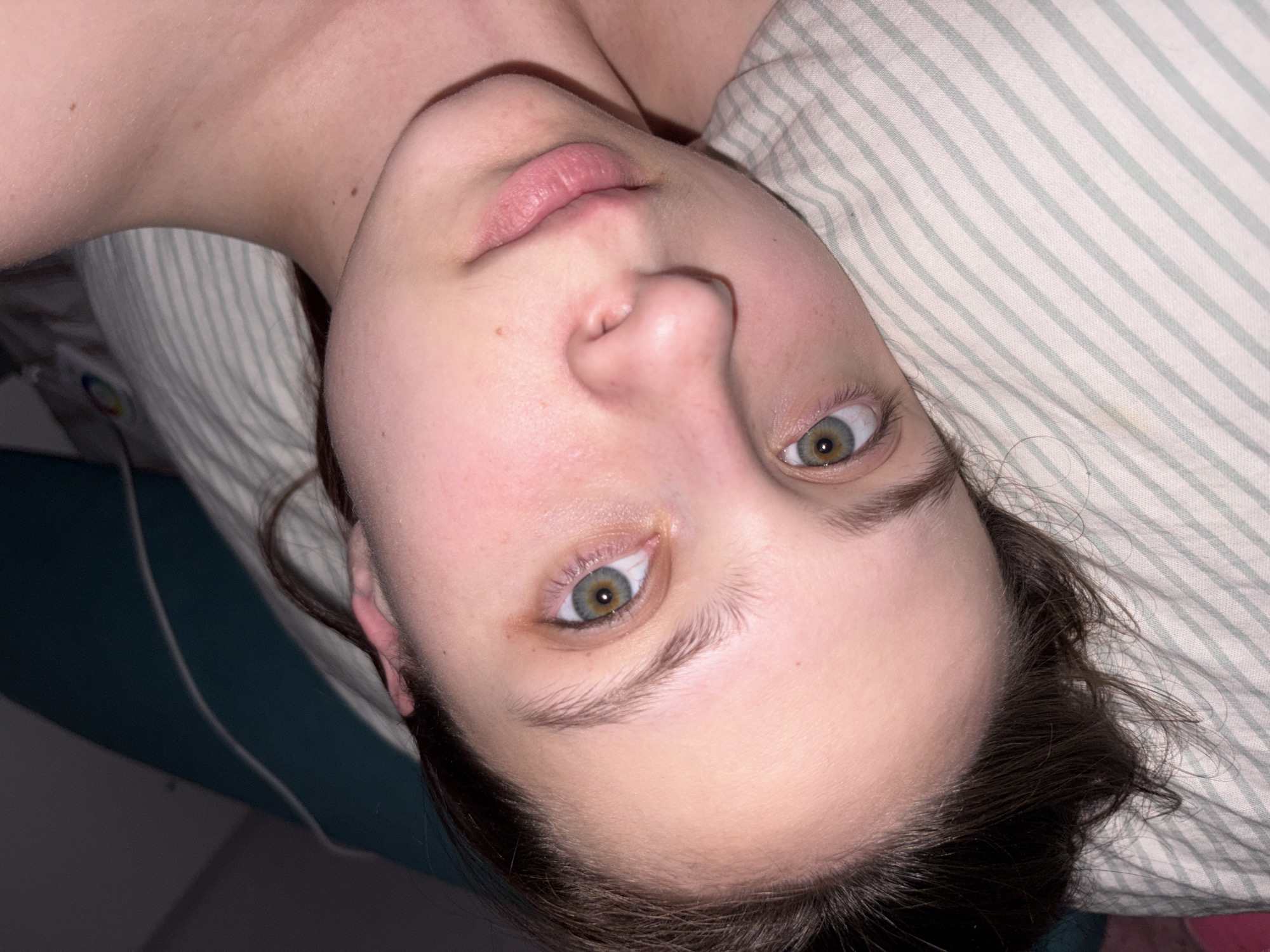 A close-up photograph depicts a person lying on their side, possibly in bed, with their head resting on a striped pillow. The person has fair skin, brown hair, and greenish-hazel eyes. The focus is on their face, capturing details such as their eyebrows, eyelashes, nose, and lips. Part of their shoulder and neck are also visible. The background includes a teal wall and a white charging cable. There is no information available to determine the city of Прикамский, Russia.