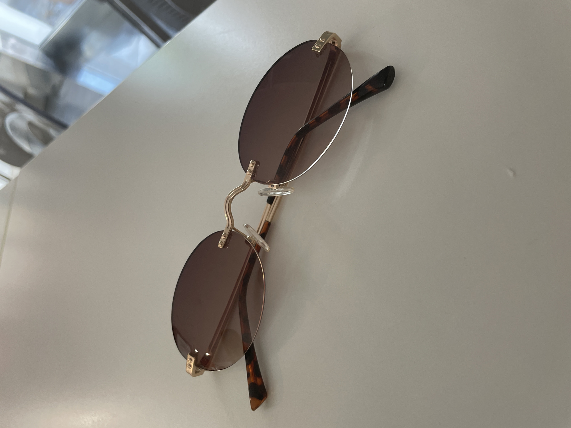 An overhead, medium shot shows a pair of rimless oval sunglasses with brown lenses and gold frames resting on a white, textured surface. The sunglasses are positioned diagonally across the frame, with the left lens closer to the viewer and the right lens further away. The temples of the sunglasses have a tortoiseshell pattern and are folded inwards. The background is out of focus, but appears to be a reflective surface with indistinct shapes and colors. The overall lighting is bright and diffused.