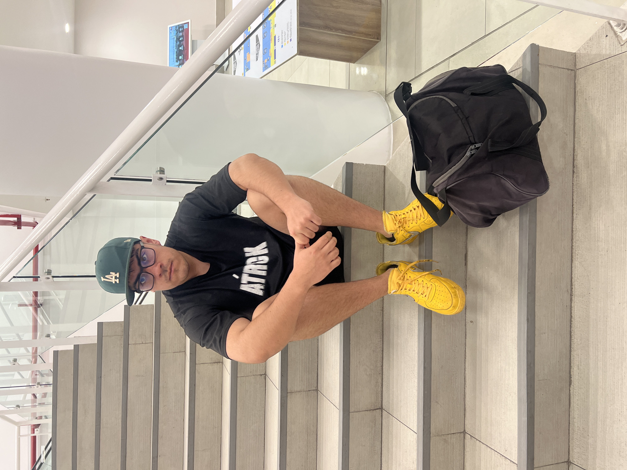 A person sits on a set of concrete stairs inside a modern building. The person is wearing a black t-shirt with a white logo, black shorts, a dark green baseball cap with an "LA" logo, and yellow sneakers. The person is fair-skinned, wearing glasses, and has short brown hair. They are positioned in the center of the frame, angled slightly toward the upper left corner, with their legs crossed and hands resting on their knees.
To the right of the person is a black backpack sitting on the upper landing of the staircase. The background includes a white wall with a glass railing, metal supports, and a display board with signage and informational graphics. The staircase is concrete, with metal edgings. The building appears to be located in an urban area, but no definitive city landmarks are visible. The lighting is bright and seems to be artificial. It is unknown whether this image was taken in the city of Arica, Chile as no visual clues can confirm this statement.