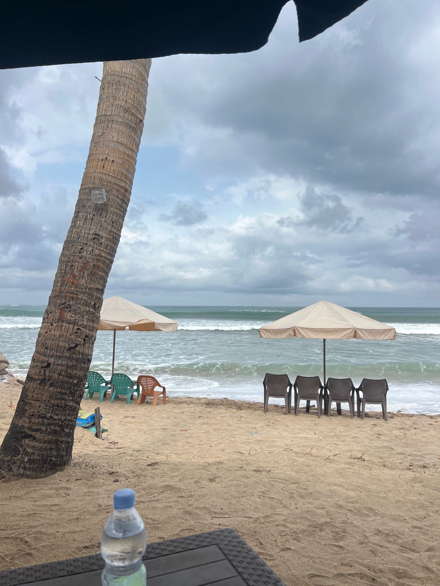 The image depicts a beach scene in Kuta, Indonesia, under a cloudy sky. In the foreground, a dark, textured surface, likely a table, is visible, with a bottle of water on it. A large palm tree trunk dominates the left side of the frame. Further back, on the sandy beach, are two beach umbrellas, one beige and situated near a group of three green plastic chairs and one orange plastic chair, and another beige umbrella closer to the right, with four dark plastic chairs lined up in front of it. The ocean is visible in the background with moderate waves. The overall atmosphere is tranquil and suggestive of a quiet day at the beach.