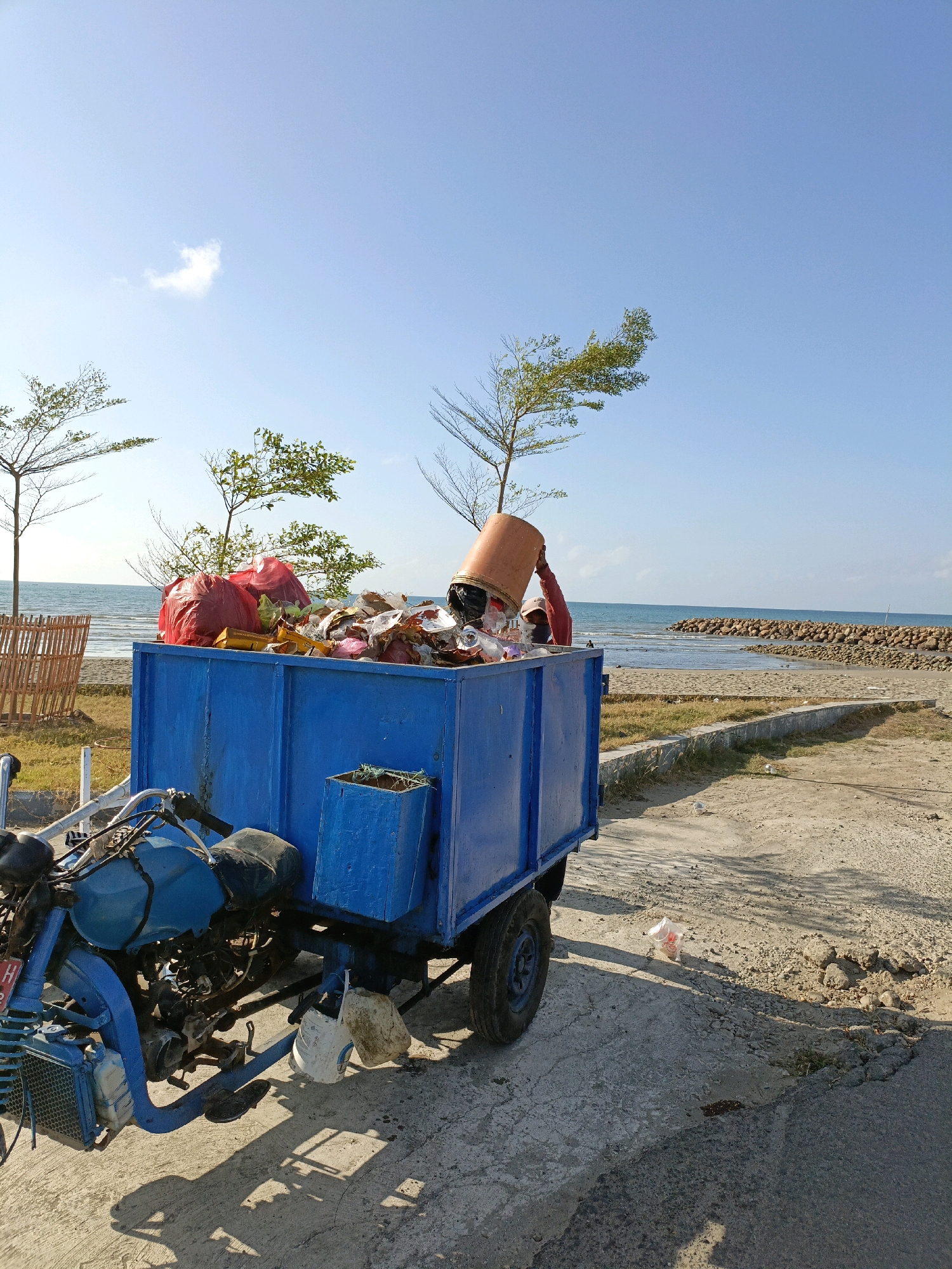A blue, three-wheeled vehicle with a large, blue trailer is parked on a dirt and asphalt road near the coast. The trailer is filled with various types of trash, including red garbage bags, plastic bottles, and a large orange bucket being held by a person. The person's arm and torso are visible as they reach into the trailer. In the background, the ocean stretches out to the horizon, with a rock jetty extending into the water. Several sparse trees with green foliage dot the landscape between the road and the beach. The sky is clear and blue, with a few small clouds. The scene appears to be in Bulukumba, Indonesia, likely a coastal area used for waste disposal or collection.