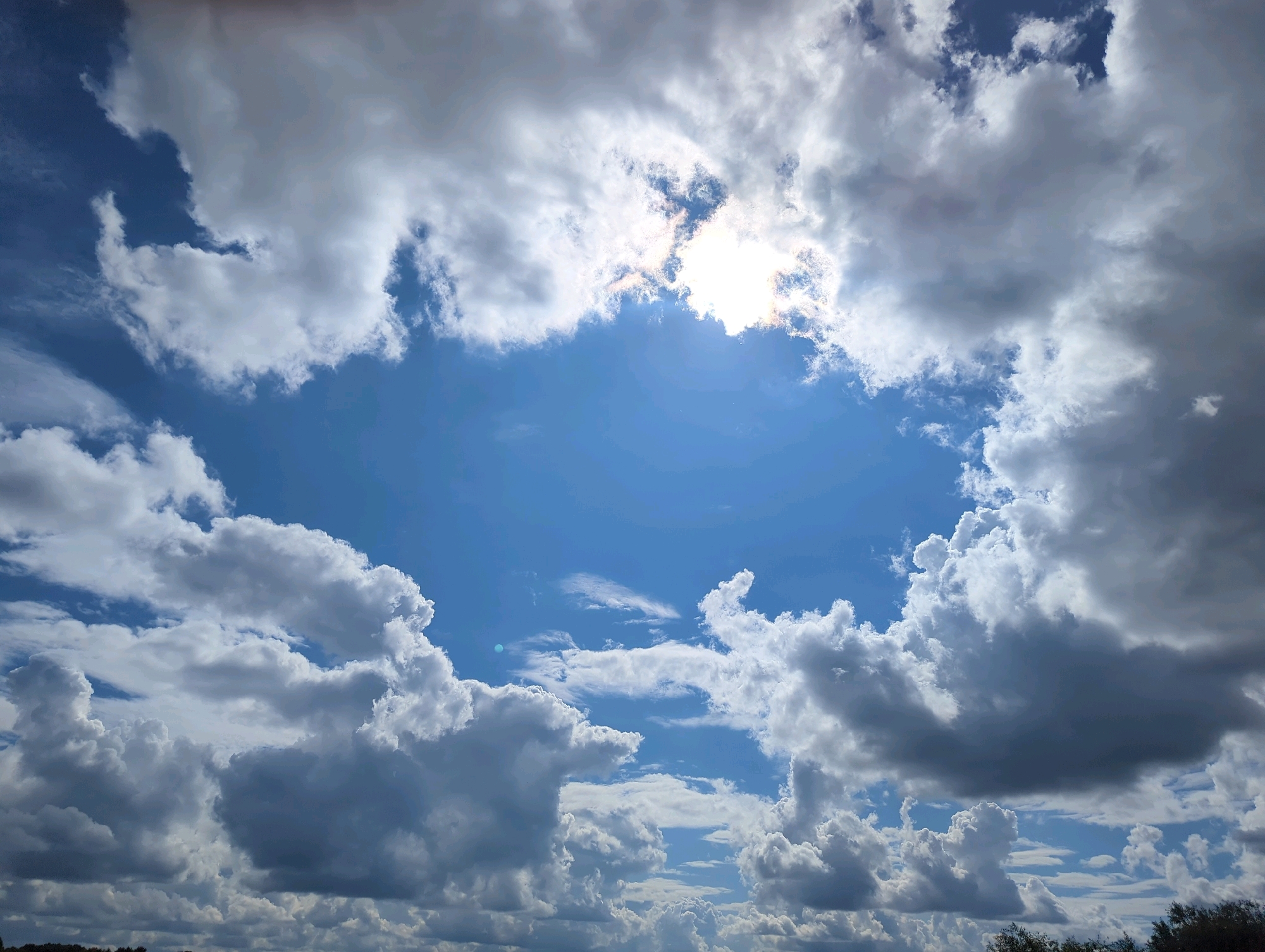 This is a photograph of a partly cloudy sky, taken from ground level looking upwards. The sun is visible, partially obscured by clouds, creating a bright, hazy glow. The clouds are a mix of white and dark grey, indicating varying densities and potentially different weather conditions. The sky itself is a vibrant blue. In the distance, along the horizon, a faint line of trees is visible, suggesting an outdoor setting possibly on the outskirts of a city. There are no people, animals, or structures in the immediate foreground or midground. The image does not contain any specific landmarks or indicators that would place it in the city of Vladimir, Russia.