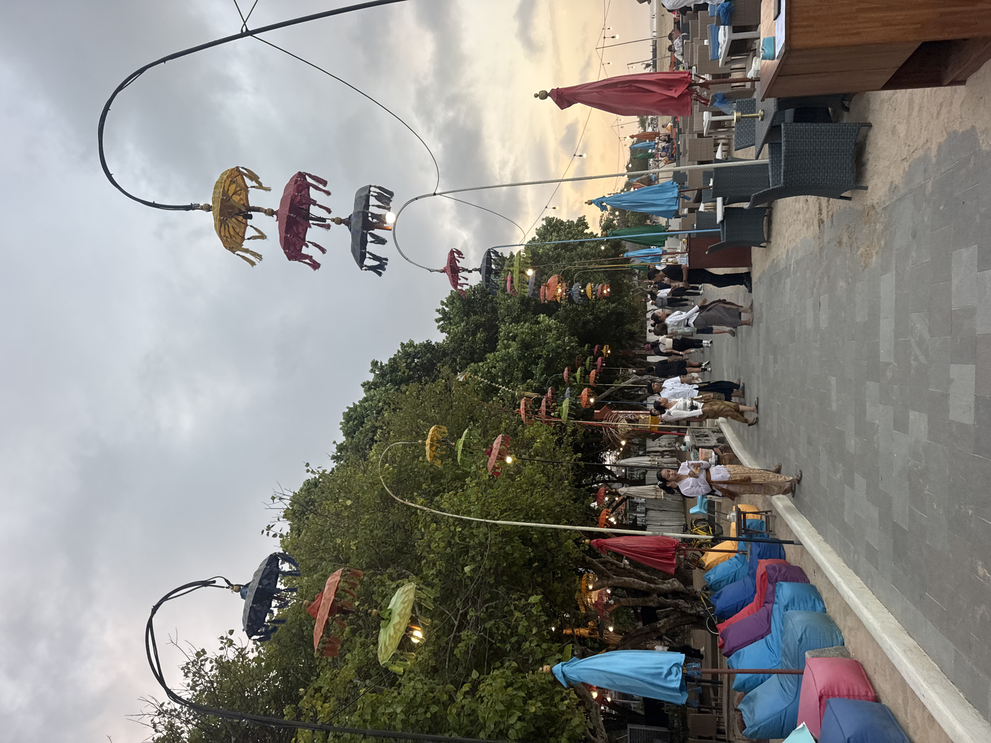 The image depicts a beachfront promenade in Kuta Selatan, Indonesia, likely at sunset. Colorful, traditional Balinese umbrellas, often referred to as "payung" or "umbrellas," are suspended from curved poles along the walkway, creating a festive and decorative atmosphere. These umbrellas are arranged in tiers and feature vibrant colors such as red, yellow, green, and black, with tasselled edges. Some of the umbrellas are illuminated by visible light bulbs.
The promenade is paved with gray square tiles and runs alongside a sandy beach. To the left of the walkway, a line of large, comfortable beanbag chairs in various bright colors (blue, purple, red, orange) are set up, indicating a relaxed seating area, possibly for beachgoers or cafe patrons. Further back, trees provide a backdrop of lush greenery.
On the promenade itself, several people are visible. Some are walking, while others appear to be socializing or working. Two women in traditional Balinese attire, characterized by sarongs and light-colored tops, are standing near the beanbag chairs, one of them holding a small object. In the background, a row of outdoor seating with tables and chairs suggests a restaurant or cafe area, with more people seated and dining. Umbrellas, some closed and some open, are scattered throughout this dining area.
To the right of the promenade, a wooden counter or bar structure is visible. The overall scene is one of a popular, lively tourist destination, with a blend of cultural elements and leisure amenities. The sky is overcast with a hint of warm light from the setting sun visible on the horizon behind the scattered umbrellas and tables.