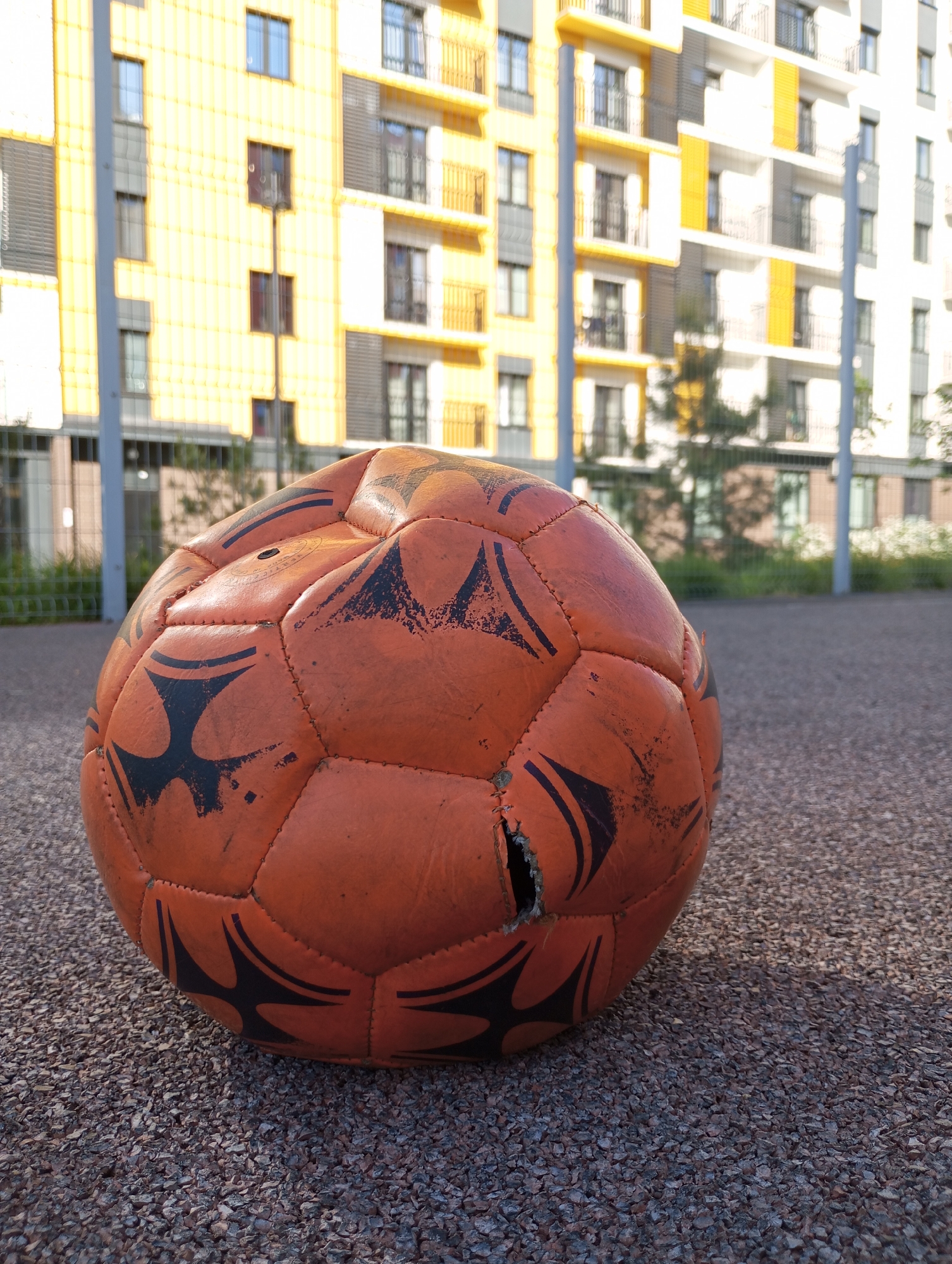 An orange, partially deflated soccer ball rests on a textured asphalt surface. The ball is visibly worn with black markings and a small tear near the center. In the background, there is a modern apartment building with yellow and white facades and multiple balconies. A section of a chain-link fence is also visible behind the ball. The setting appears to be in an urban area, possibly a playground or recreational space, located in Pervomaysky, Russia. The time is likely daytime, based on the sunlight and shadows present. There are no people visible in the image.