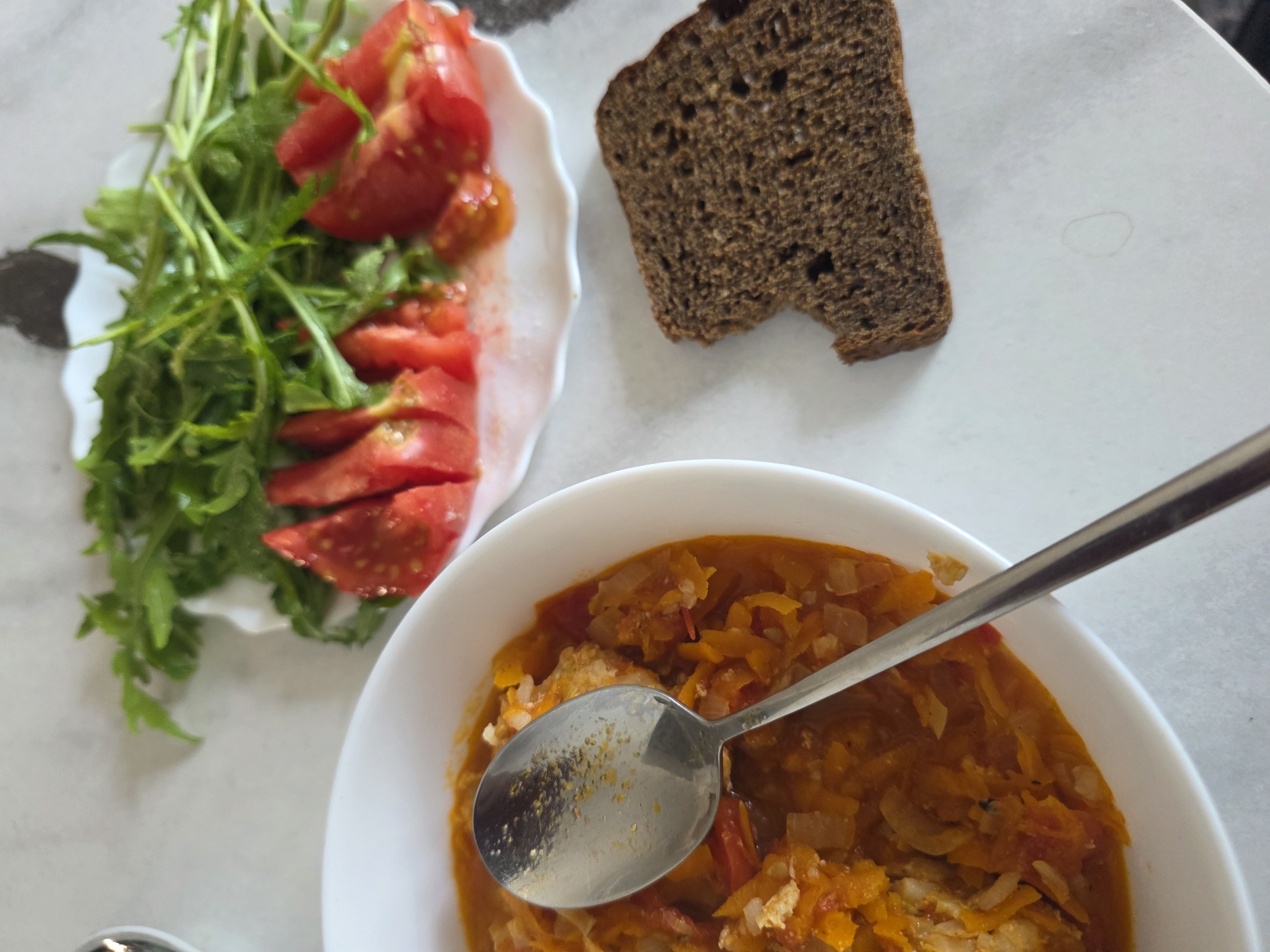 In an indoor setting, a meal is presented on a white surface. A white bowl of stew, rich in orange and red hues and containing visible pieces of vegetables and possibly fish, is prominently featured, with a silver spoon resting inside. To the right of the bowl, a slice of dark, whole-grain bread lies on the surface. Further to the right, a white plate displays a salad composed of fresh green arugula and sliced red tomatoes. The lighting suggests a bright, natural source, casting subtle shadows on the table.