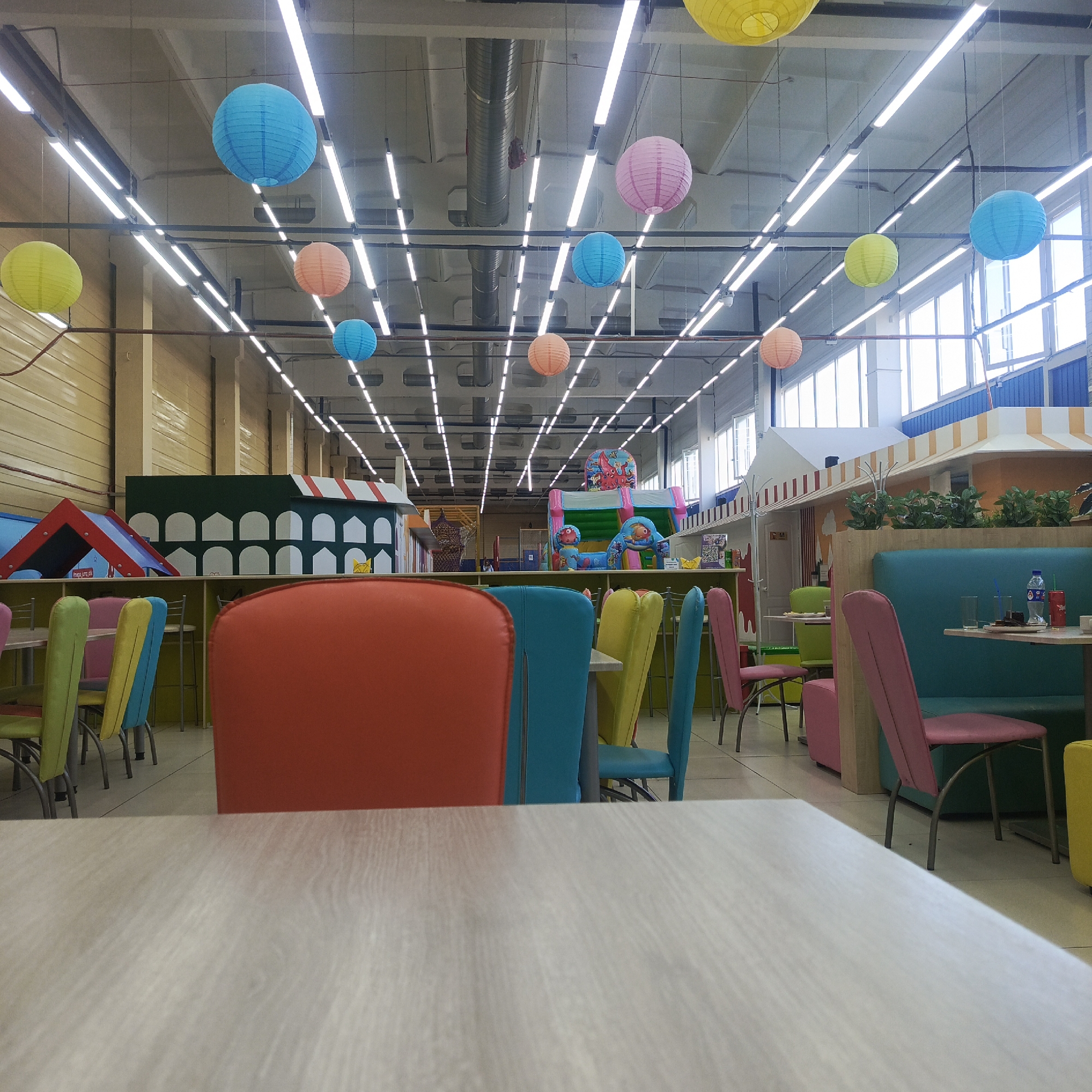 The image shows the interior of a children's entertainment center, possibly a restaurant or cafe associated with a play area, located in Chaikovsky, Russia. The foreground features a light-colored tabletop with colorful chairs in shades of red, blue, green, yellow, and pink surrounding it. Behind the chairs, there is a partially visible play structure featuring a green facade with white arched windows and a red-and-white striped roof. In the background, an inflatable play area with cartoon figures is visible. Further back, there are stylized building facades. Overhead, the ceiling is a grid of white fluorescent lights interspersed with colored paper lanterns. There are also windows providing natural light. A small number of people is visible towards the back of the space. There's also a booth-style seating area in the background.