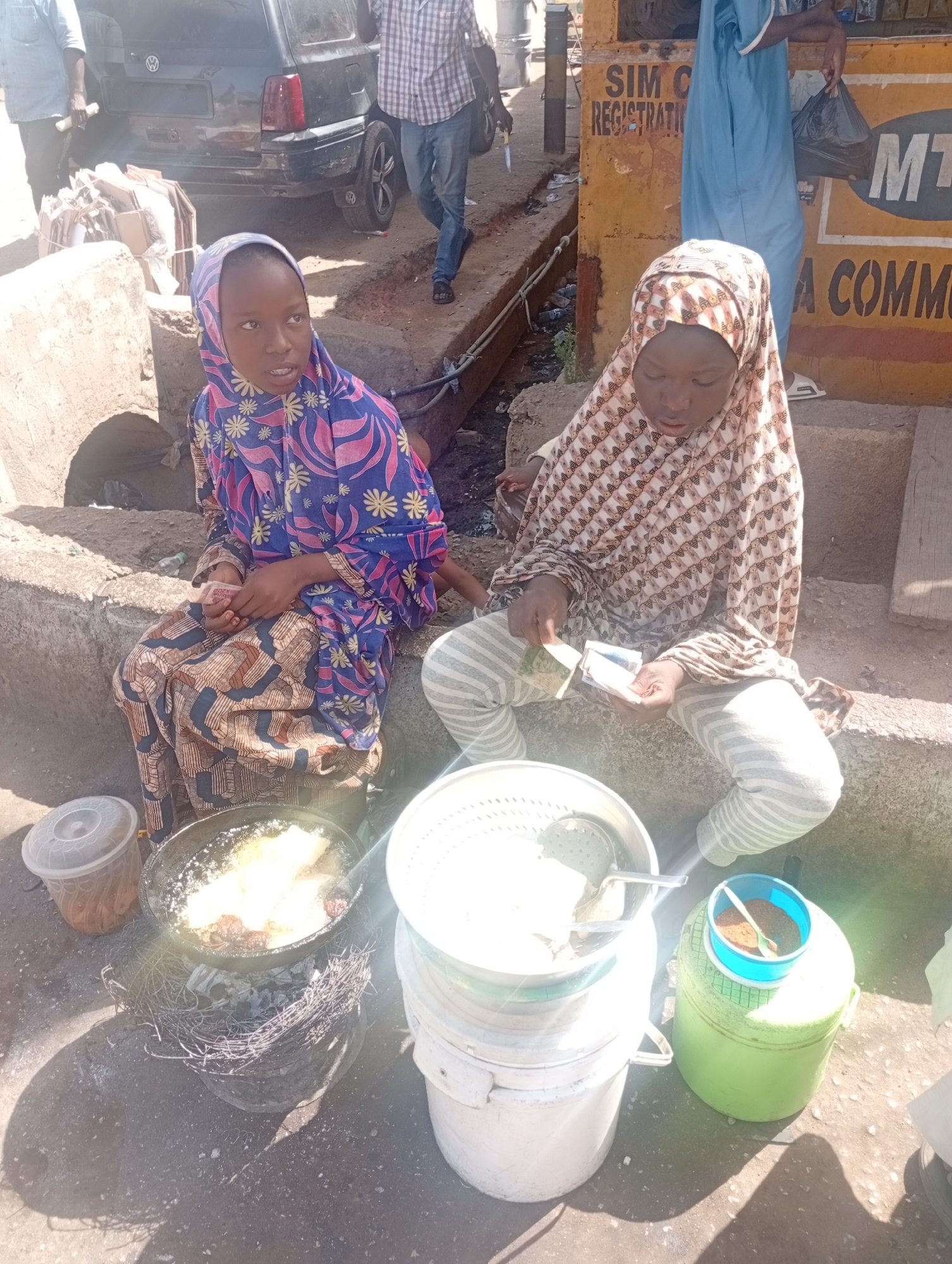 Two young girls are seated on a concrete ledge on the street, likely in Jos, Nigeria, implied by the presence of a sign reading "SIM C REGISTRATION" and "MTN A COMMUN". One girl wears a blue and pink floral head covering and dress. The other girl is clad in a brown and white patterned hijab and striped pants. They are positioned next to a cooking setup with a frying pan over a charcoal brazier and a stack of white buckets containing a colander. The girl in the brown and white hijab is holding and counting Nigerian currency. In the background is a black car, pedestrians, and parts of buildings, implying a street scene.