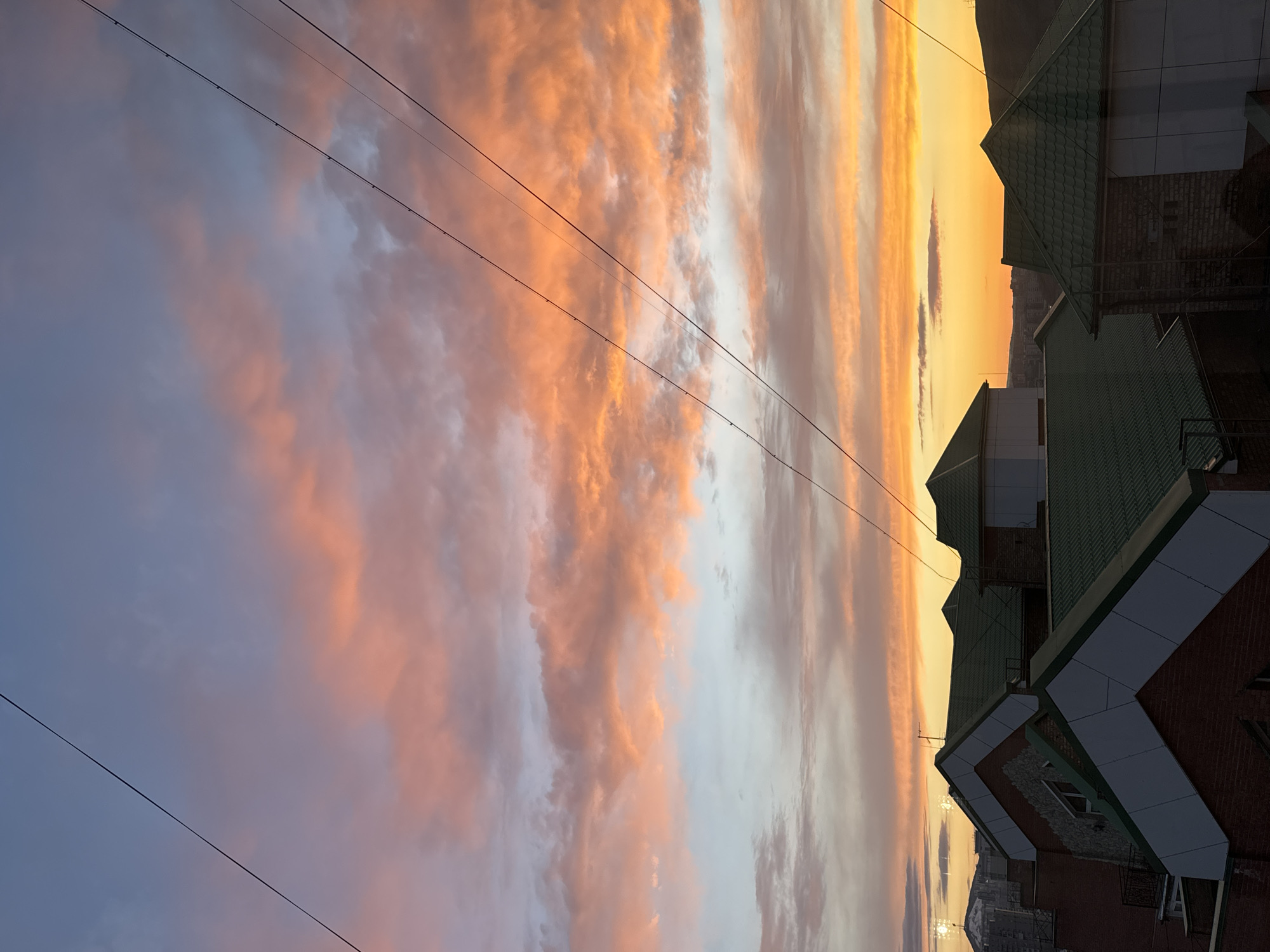 The image shows a sunset sky with pink and orange clouds, and buildings in Krasnoyarsk, Russia, with green roofs and brick facades. Several power lines are running across the sky. In the distance, the mountains outside Krasnoyarsk are visible.