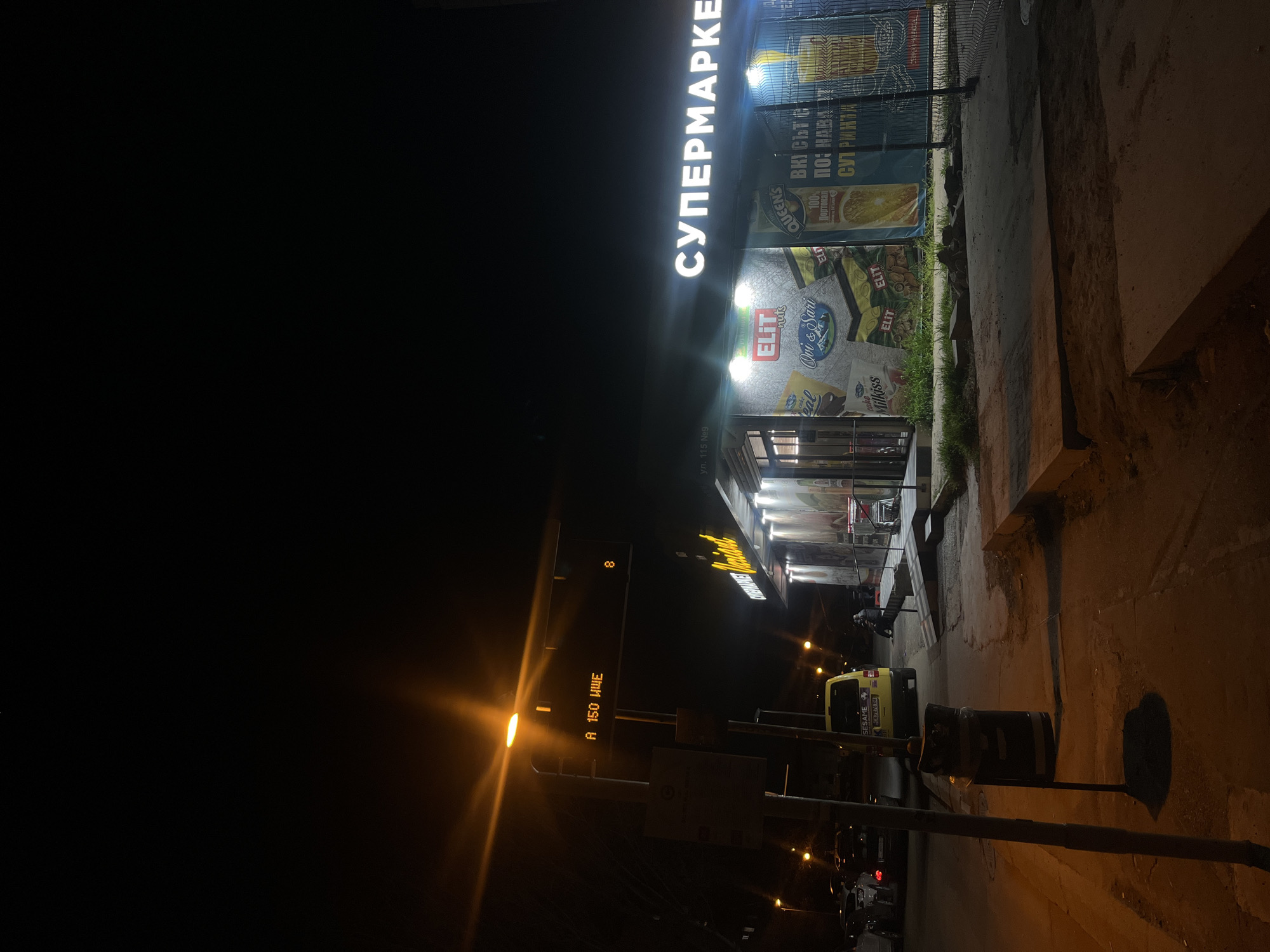 The image depicts a nighttime scene in Sofia, Bulgaria, showing a street view with a supermarket. The supermarket's name is brightly lit in Cyrillic letters, translating to "SUPERMARKET". Advertisements for food products line the exterior of the store. A road sign displays "A 150 ИЩЕ", likely indicating a route or location, and "ул. 115 №9", potentially identifying the street address as 115, number 9.
On the street, a yellow vehicle with "SESAME" branding is parked. A trash bin stands near the curb. Further down the street, there are other parked cars and possible pedestrians. The scene is illuminated by streetlights, creating a mix of shadows and artificial light.