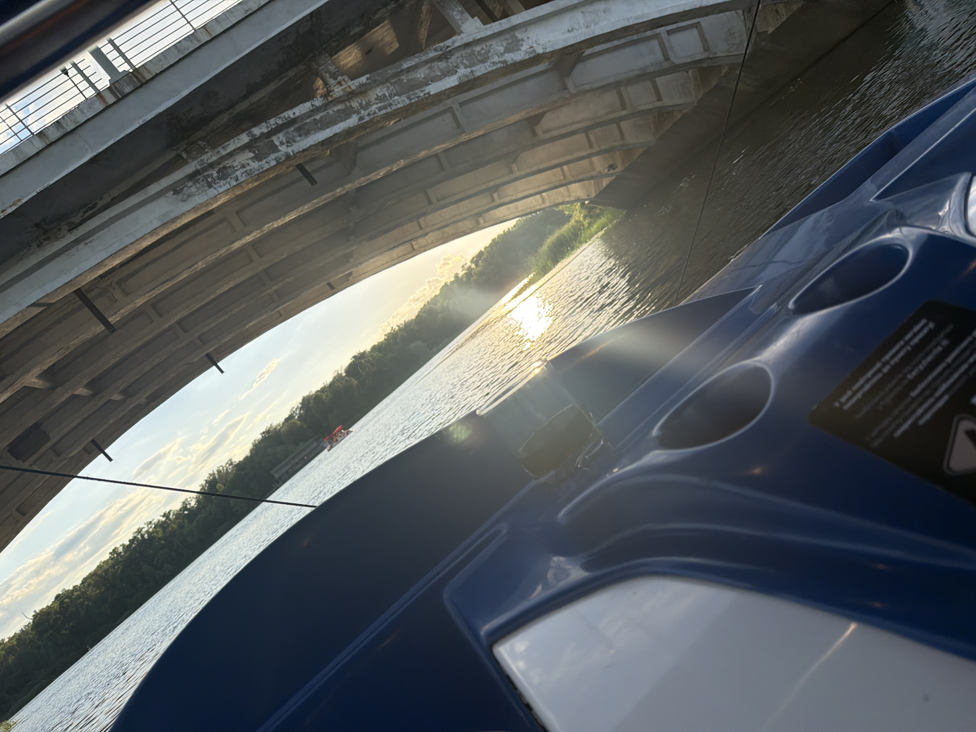 This media file depicts a serene scene on a body of water, possibly a river, as viewed from the perspective of a boat. A large concrete bridge with visible support beams dominates the upper portion of the frame, casting a shadow over the water. The sun is setting, as indicated by the warm golden light reflecting off the water's surface and the soft, hazy glow in the sky. The distant shoreline is lined with lush green trees, suggesting a natural, possibly rural or park-like setting.
The foreground is occupied by the blue and white structure of a boat, possibly a pedal boat or a similar recreational craft. The smooth, curved lines of the boat's hull are visible, along with a few circular indentations that might be cup holders. A black warning label with some text and a triangular symbol is affixed to the boat's surface. The text on the label is small and partially obscured, but some legible words include "and", "to", "for", and possibly safety instructions.
The overall mood is peaceful and tranquil, with the gentle ripples on the water and the soft sunset light contributing to a sense of calm. The presence of the bridge implies an urban or suburban setting nearby, yet the natural elements of the water and trees create a contrast. The time of day is clearly late afternoon or early evening, coinciding with the golden hour. There are no visible people in the immediate foreground, but a small red and white boat is visible in the distance on the water, suggesting other people are enjoying the waterway. The location context of Kyiv, Ukraine, adds a layer of geographical specificity to this picturesque scene.