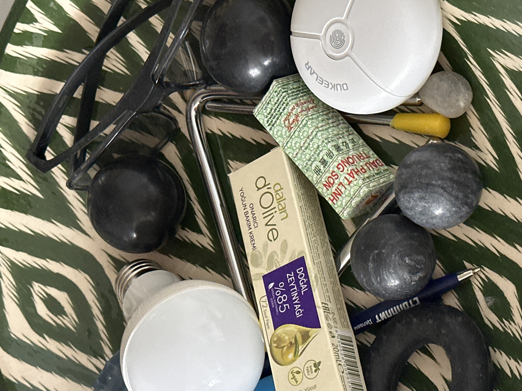 The image displays a collection of miscellaneous objects arranged on a green and white patterned surface. The objects include a white LED light bulb, several dark spherical objects that appear to be stones or marbles, a box of "dalan Olive" skincare product with Turkish writing, a roll of what appears to be traditional medicine in a green patterned wrapper with Asian writing, a blue pen, a black ring-shaped object, a yellow pointed object, and a white circular device with the word "DUKEELAR" on it and a fingerprint icon. Also visible are a pair of black eyeglasses and a metallic handle. The lighting suggests an indoor environment, possibly a tabletop or shelf. There is no indication of the city of Nevinnomyssk, Russia.
