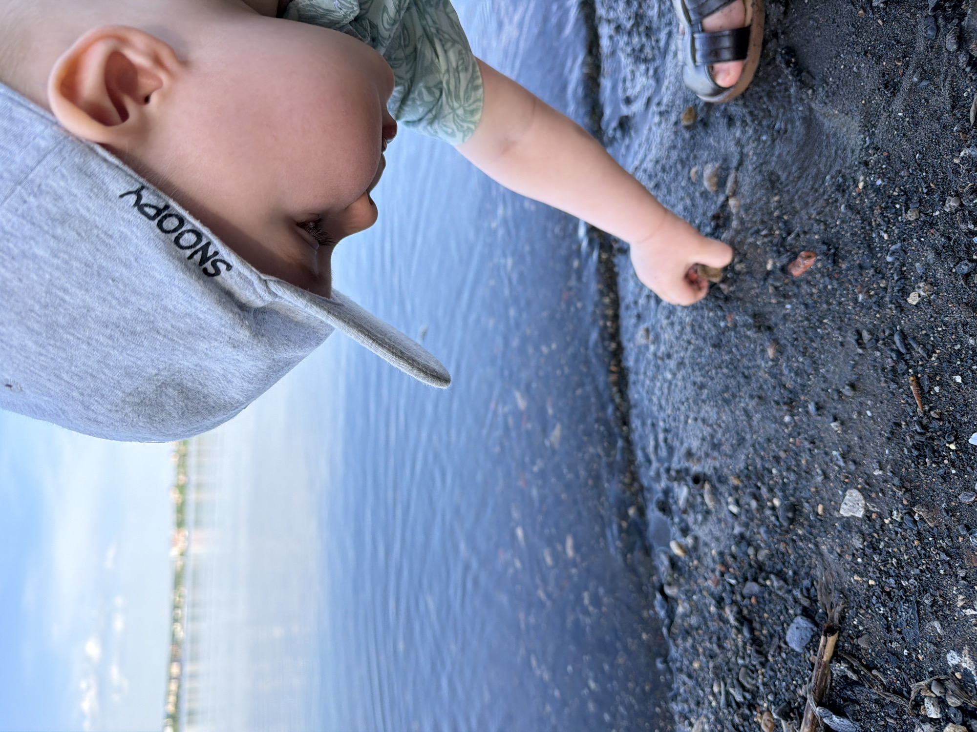 A young child wearing a grey "SNOOPY" hat and a green printed shirt is standing at the edge of a body of water, possibly a lake or river. The child is reaching down towards the dark, gravelly shoreline with their right hand, as if picking up a small rock or object. Part of a sandal-clad foot is visible in the upper right of the image. The water is blue-grey and calm, and across the water in the distance is a cityscape, which could potentially be Votkinsk, Russia. The sky is light blue with some cloud cover.