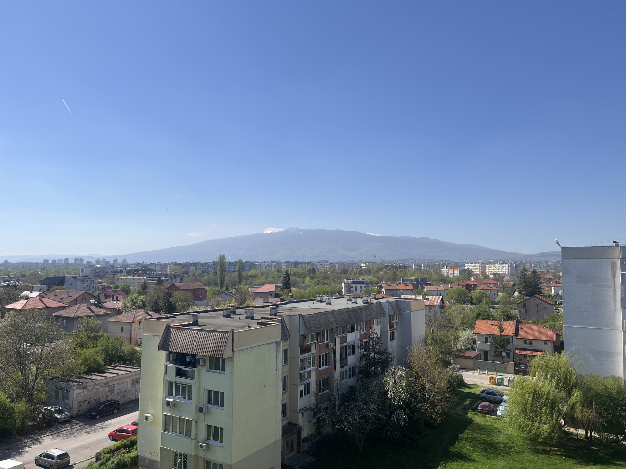 This high-angle, wide shot captures a bright, sunny urban landscape with a majestic snow-capped mountain range in the distant background. The scene is set in what appears to be a residential area of Sofia, Bulgaria, identifiable by the characteristic architecture and the iconic Vitosha Mountain.
In the foreground, a multi-story apartment building with a light yellow facade dominates the lower right, featuring numerous windows, balconies, and air conditioning units. Its roof is flat with visible vents. To its left, another, older apartment block with a dark, corrugated roof extends, showing signs of wear and multiple balconies. A dense array of trees, some with light green spring foliage or white blossoms, fills the spaces between buildings and lines the streets. Green grassy areas are prominent, suggesting springtime.
A street runs through the scene, with several cars parked on the side and in small lots, including red, silver, and dark-colored vehicles. In the lower-left quadrant, a smaller, dilapidated building with graffiti on its walls is visible, bearing the Bulgarian text "АВТОМИВКА" (AVTOMIVKA), meaning "Car Wash." Other legible text includes "ЦСК" (CSK or TSK) on the side of the light yellow building.
Beyond the immediate neighborhood, a sprawling cityscape extends to the horizon, characterized by a mix of houses with red tile roofs and more distant multi-story buildings. In the far background, the impressive Vitosha Mountain stands tall, its upper slopes still covered in patches of snow under a clear blue sky. A faint airplane contrail streaks across the upper left sky, and a few small birds are seen closer to the horizon. On the right edge of the image, a tall, white cylindrical structure (possibly a water tower or part of a building) rises above the rooftops.
The weather is clear and sunny, indicative of a pleasant day, likely late morning or early afternoon, given the bright illumination and distinct shadows. The overall mood is peaceful and serene, showcasing typical daily life in a European city framed by nature.