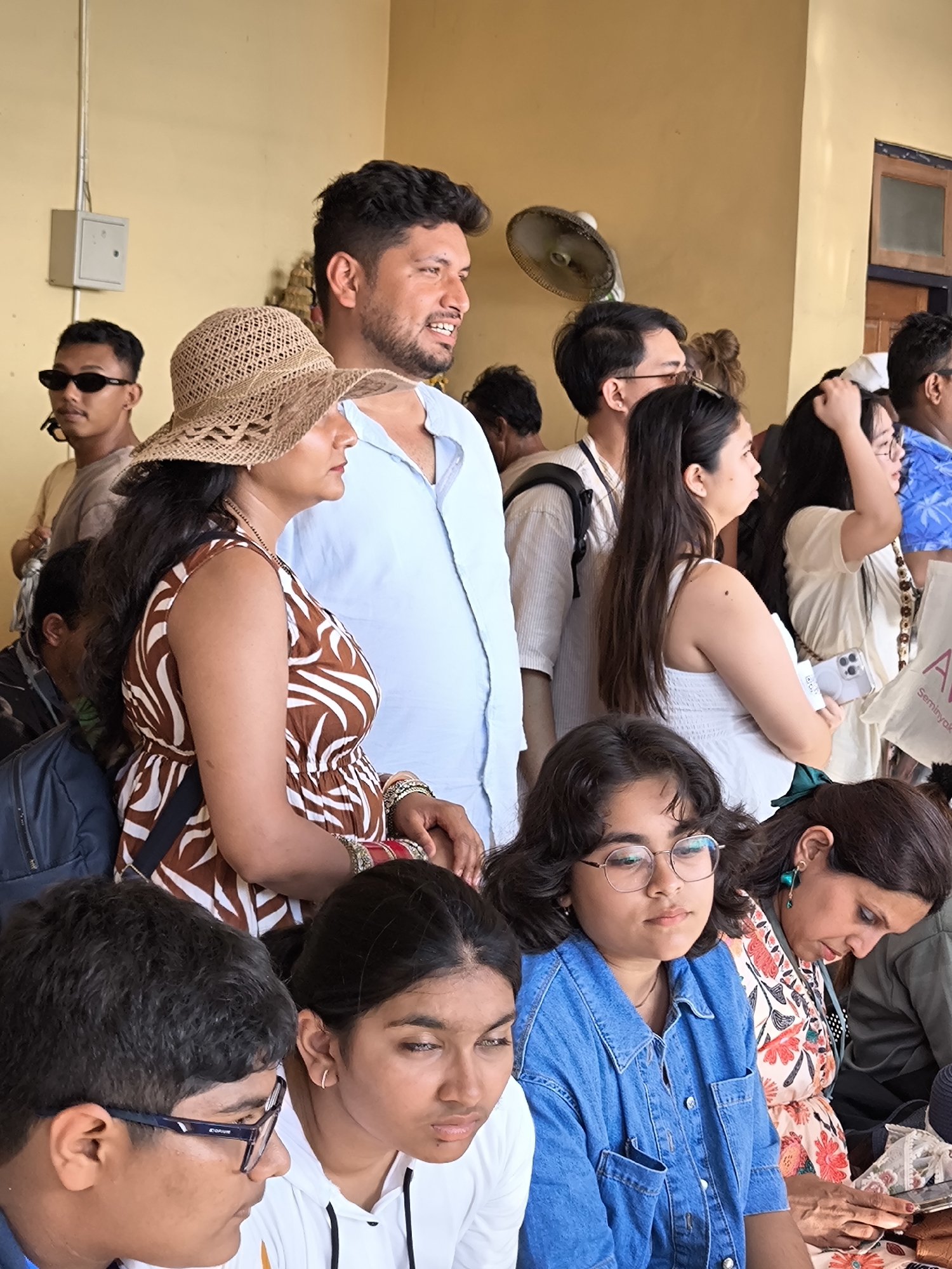 A daytime medium shot depicts a diverse group of individuals in an indoor setting. An overhead electric fan is visible against a pale yellow wall, and a small grey electrical box is mounted high on the wall to the left.
Several individuals are visible, including:
- A woman in a brown and white patterned dress and straw hat.
- A man in a light blue shirt.
- A woman in a white dress.
- A girl in a denim jacket wearing glasses.
They are arranged in a crowded space, some individuals are seated, and others stand. They do not interact with each other in any way, and it seems like a lot of people are observing something outside of the view of the camera. A shopping bag with text on it that reads "A Seminyak" is also visible.
The location of the photo is Nusa Penida, Indonesia.