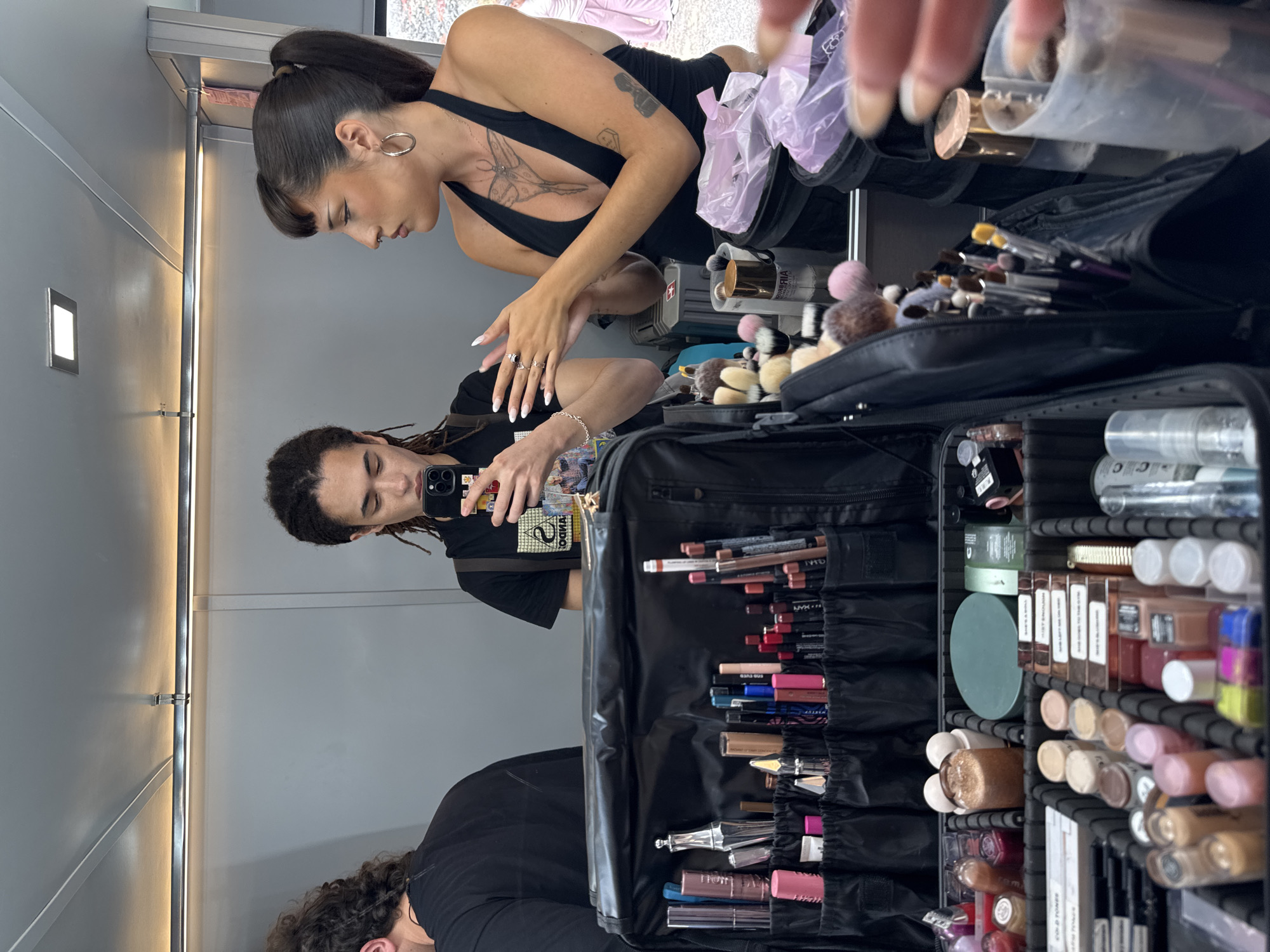 The image shows the interior of what appears to be a dressing room or makeup area, potentially within a film or photography studio in Tinajo, Spain. A woman with dark hair in a high ponytail and a black top is visible in the reflection of a mirror; she has a tattoo on her chest. Another individual with dreadlocks is holding a smartphone and capturing the scene, reflected in the same mirror. A large, black makeup case is open, revealing a wide array of makeup products including lipsticks, eyeliners, mascaras, and other cosmetic items. Additionally, makeup brushes and accessories are also present. The backdrop includes a silver-toned wall.