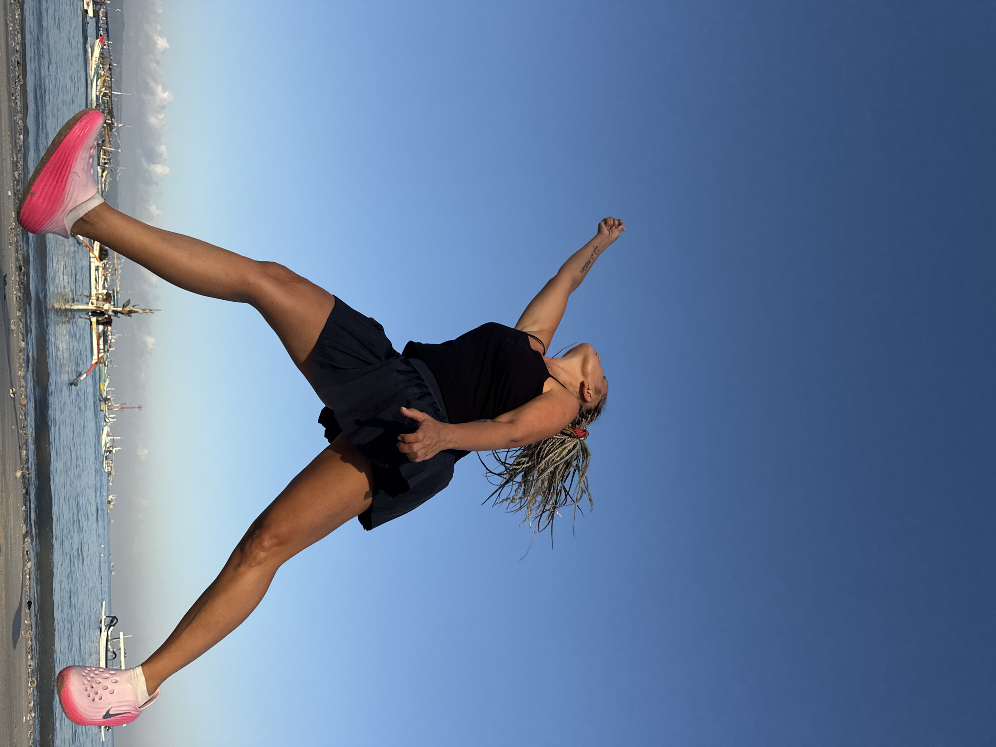 A woman with braided blonde hair is captured mid-jump against a clear blue sky, her body angled to the right. She wears a black tank top and a dark skirt, with bright pink sneakers on her feet. Her left arm is raised and extended upwards, and her head is tilted back. In the background, the ocean is dotted with numerous small boats, characteristic of Kuta, Indonesia. The water is calm, and a few clouds are visible on the horizon. She appears to be on a sandy or rocky shoreline.