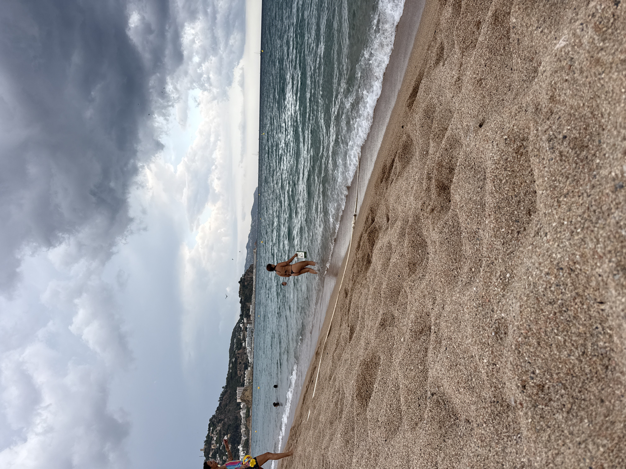 The image depicts a wide sandy beach in Blanes, Spain, with the Mediterranean Sea on the right and a town nestled on a hill in the background on the left. The sky is overcast with dramatic, dark clouds, suggesting impending rain. In the foreground, the textured sand stretches across the frame, with a rope demarcating the edge of the beach.
Several people are visible in the water. On the left, a person in a multi-colored top and dark shorts stands on the sand, holding a yellow object. Further in the water, two individuals are swimming. On the right, a woman wearing a dark bikini walks in the water and holds a light-colored object, possibly a book or a small cooler. The sea has moderate waves breaking on the shore.
In the background, the coastal town of Blanes is visible, with buildings clustered on the hillside. Several yellow buoys are scattered in the water, and a few sailboats are on the horizon. The overall scene captures a moment on a beach before or during a change in weather.