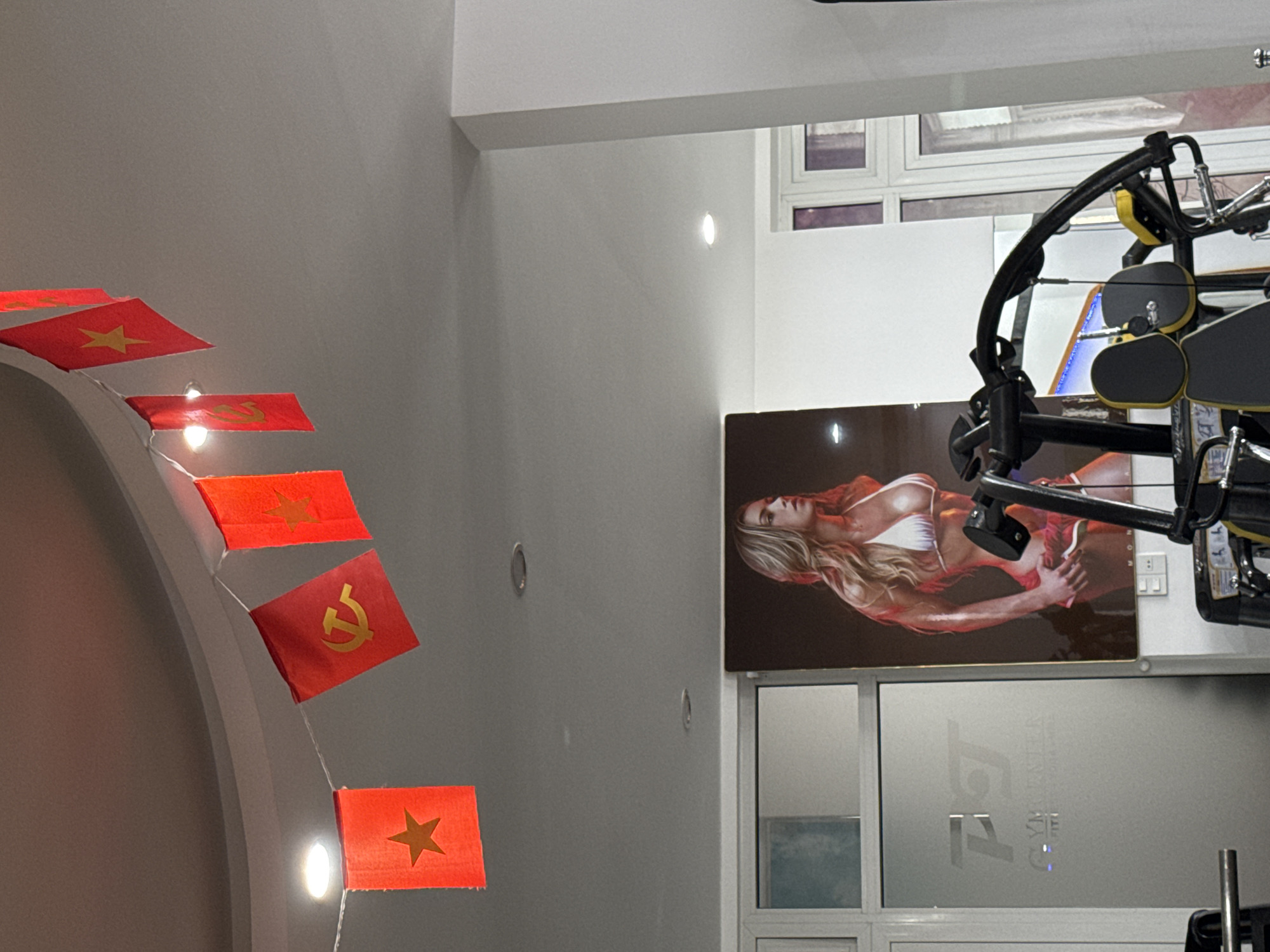 This image captures an indoor scene, likely within a gym or fitness center in Da Lat, Vietnam. The setting features light-colored walls and ceiling, illuminated by several recessed circular lights and small bulbs integrated into a string of decorative flags.
The subjects include a large poster displaying a fair-skinned woman with long, blonde hair, wearing a white bikini top, and appearing muscular. She is depicted posing with one arm behind her back. No other people are visible in the frame.
Prominent objects include a string of small red flags draped along an arched architectural feature on the left. Some flags bear a yellow five-pointed star, resembling the flag of Vietnam, while at least one prominently displays a yellow hammer and sickle, a symbol associated with communism. On the right side, partially visible black and yellow gym equipment, possibly a weight machine, suggests the fitness environment. Below the poster, a frosted glass panel, likely part of a door or large window, features visible text. Electrical outlets or switches are also visible on the wall behind the gym equipment.
The scene depicts a static interior, with no active event taking place. Notable details include the clear location cues provided by the Vietnamese and communist flags, reinforcing the provided context of Da Lat, Vietnam. The artificial lighting suggests it could be any time of day.
Visible text on the frosted glass panel clearly reads "GYM" and below it, "TRAINING FITNESS" or similar, with the overall logo appearing to be "TJ GYM FITNESS".