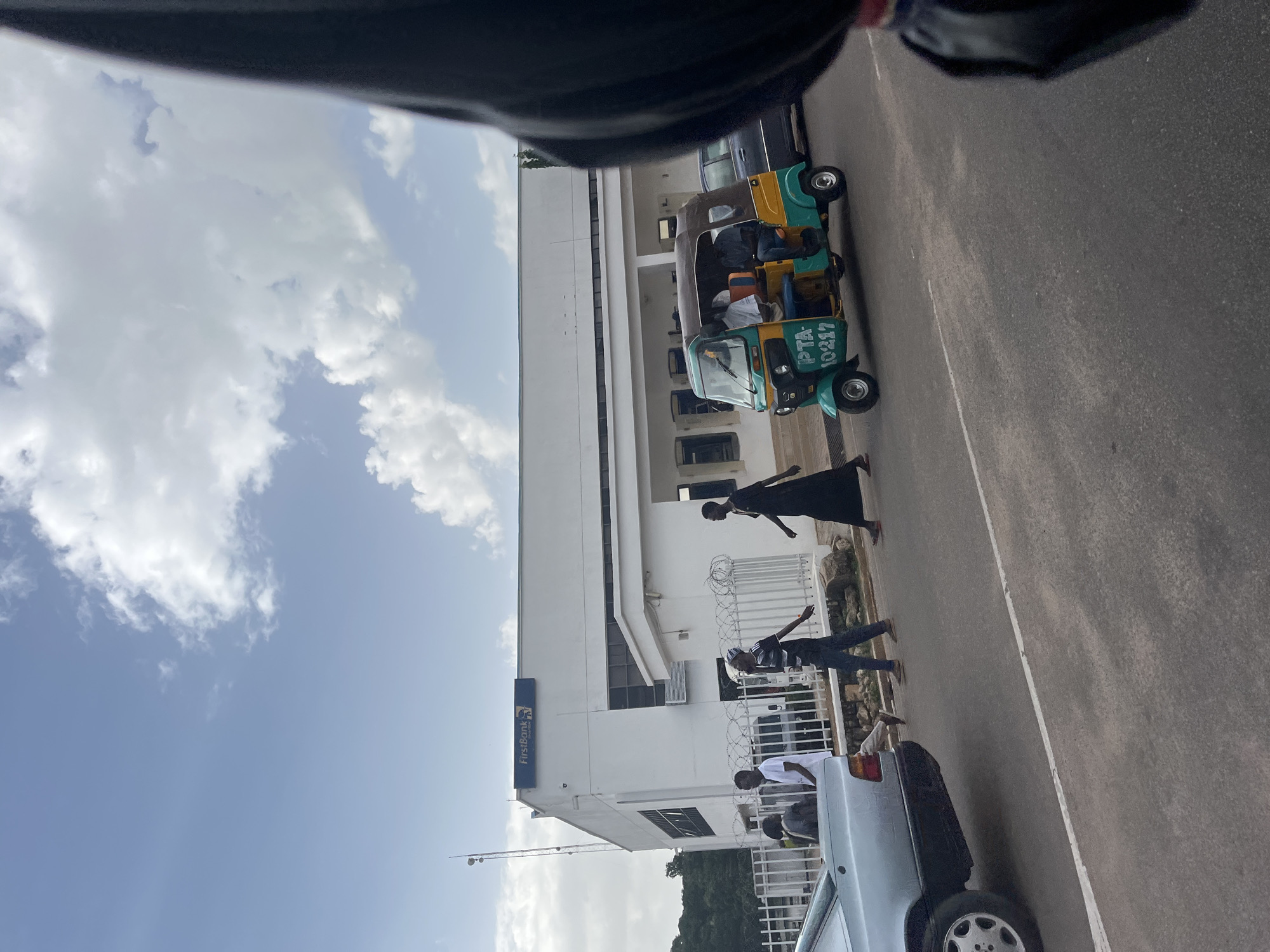 In Jos, Nigeria, a street scene unfolds under a partly cloudy sky. A white building, identified by a "FirstBank" sign, serves as the backdrop. In front of the building, a wire fence encloses the property. A light blue car is parked on the left side of the frame. Further into the scene, a colorful auto-rickshaw, painted in green and yellow with the license plate "PTA-10217," carries passengers. Several individuals are walking along the sidewalk and crossing the street. The road is paved, with a white line marking the lane. The overall impression is of a typical urban environment in Jos.