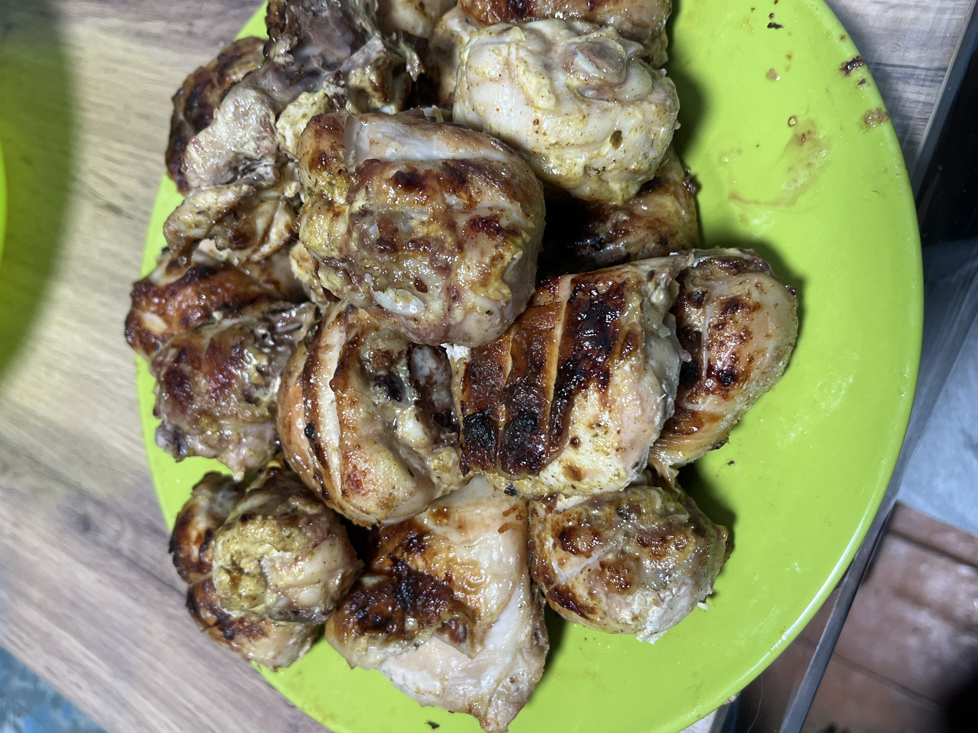 A close-up, overhead shot displays a pile of cooked chicken pieces on a bright green plate. The chicken appears to be seasoned with a yellowish spice mixture and has grill marks, indicating it was likely grilled or roasted. Some pieces show a slight charring. The plate rests on a wooden surface, possibly a table or countertop. The background is slightly blurred, with hints of a lighter green object on the left and a darker, vertical element on the right, suggesting an indoor setting. There are no people visible in the image. The lighting appears to be artificial.