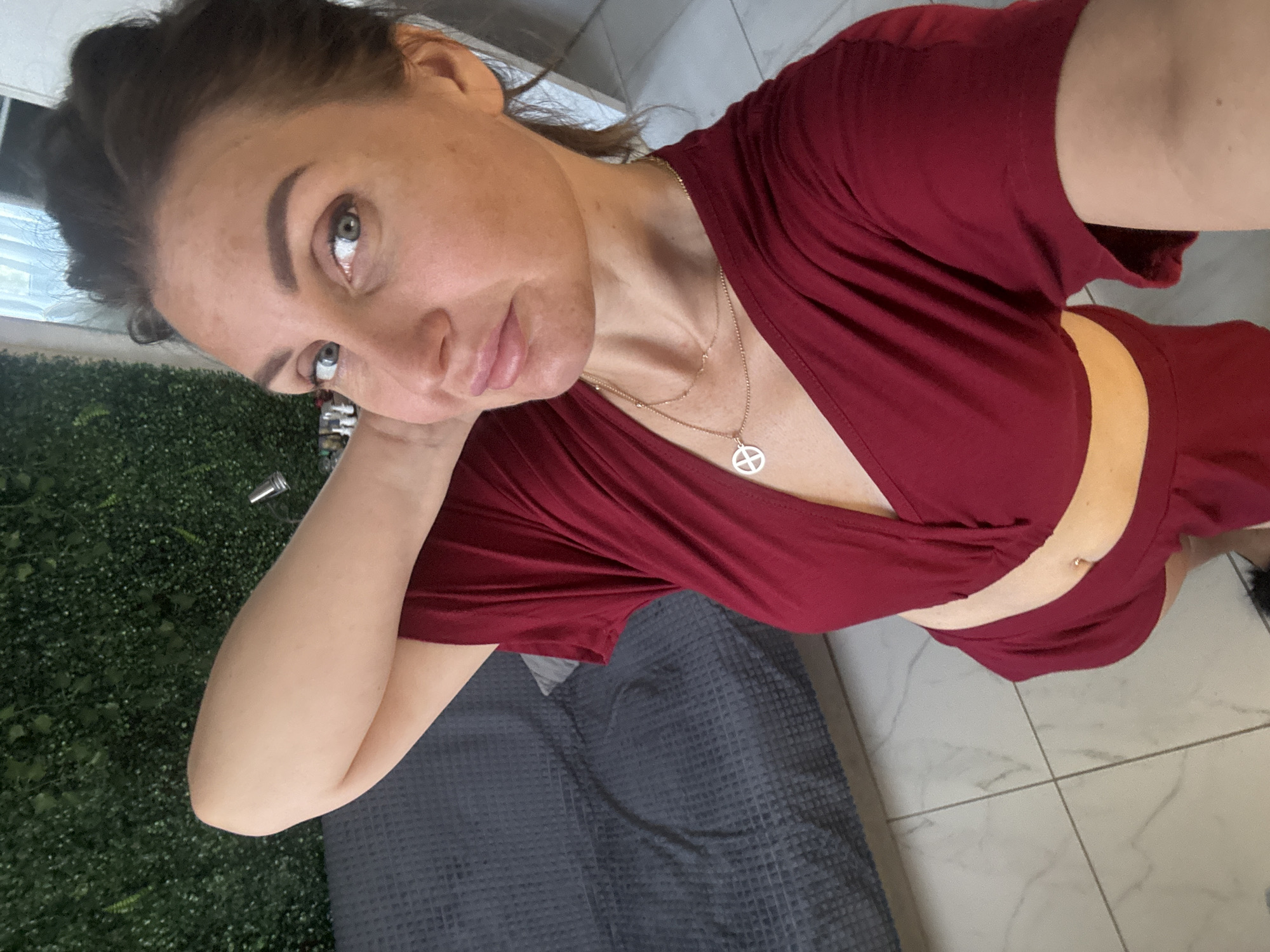 A woman is pictured in a selfie taken indoors. She is wearing a maroon v-neck top and matching shorts. A delicate gold necklace with a circular pendant is visible around her neck. She has her left arm raised, resting her hand behind her head. Her hair is pulled back in a bun, and she is looking towards the camera with a slight smile. The background shows a grey textured sofa, a wall covered in artificial green foliage, and tiled flooring. The location of Sochi, Russia is not discernible from the image.