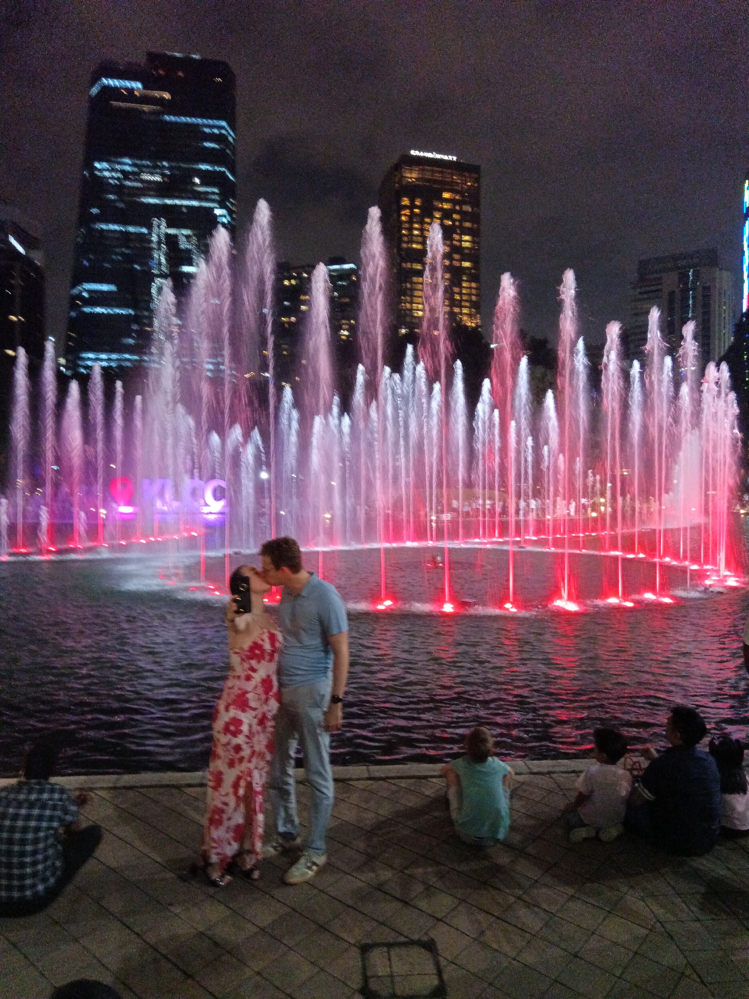 In the city of Kuala Lumpur, Malaysia, a couple kisses in front of a large, illuminated fountain display at night. The fountain features numerous jets of water, some lit with pink and red lights. In the background, tall skyscrapers, including the Grand Hyatt, are visible. The area around the fountain is paved with wood, and several people are seated on the ground, observing the fountain. One person in a plaid shirt is sitting on the left, and a family of four, including two children, are seated on the right. The overall scene depicts a romantic and lively evening at a public landmark.