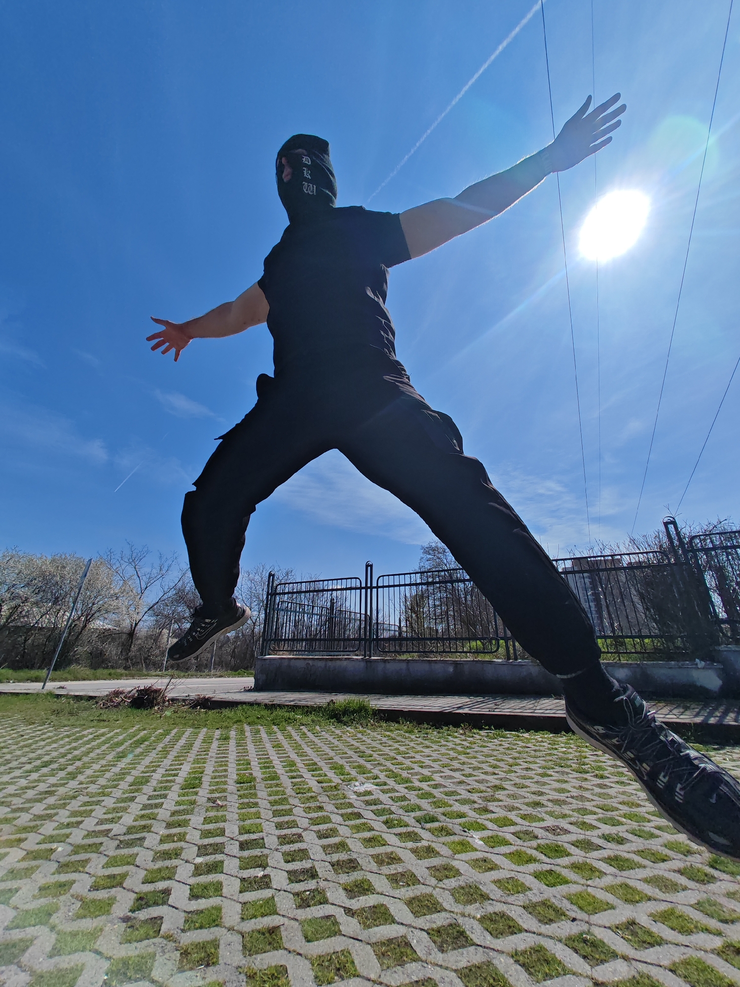 An individual is captured mid-jump against a clear blue sky in Shumen, Bulgaria. The subject is clad in a black short-sleeved t-shirt, black pants, and a black balaclava covering the face, with white lettering, possibly "DKW", visible on the left side of the head. Black athletic shoes, bearing a white Nike swoosh on the right foot, are visible on both feet. The individual's arms are extended outwards, palms open, with the left arm slightly higher. The left leg is bent at the knee, while the right leg is extended backward and downward.
The image is taken from a low angle, looking upwards. The sky is clear blue, with the bright sun prominently visible in the upper right quadrant, generating a significant lens flare. Multiple thin, dark power lines stretch horizontally across the upper portion of the frame, and a faint white contrail is visible in the upper left sky.
The ground surface beneath the individual consists of light gray concrete grid pavers with green grass growing within the open cells. In the background, to the left, are trees, some exhibiting white blossoms characteristic of early spring, alongside bare deciduous trees and a visible section of an asphalt road. To the right, a black metal fence or railing with vertical bars is situated on a concrete base. Beyond this fence, a multi-story building is partially visible through the sparse tree canopy.