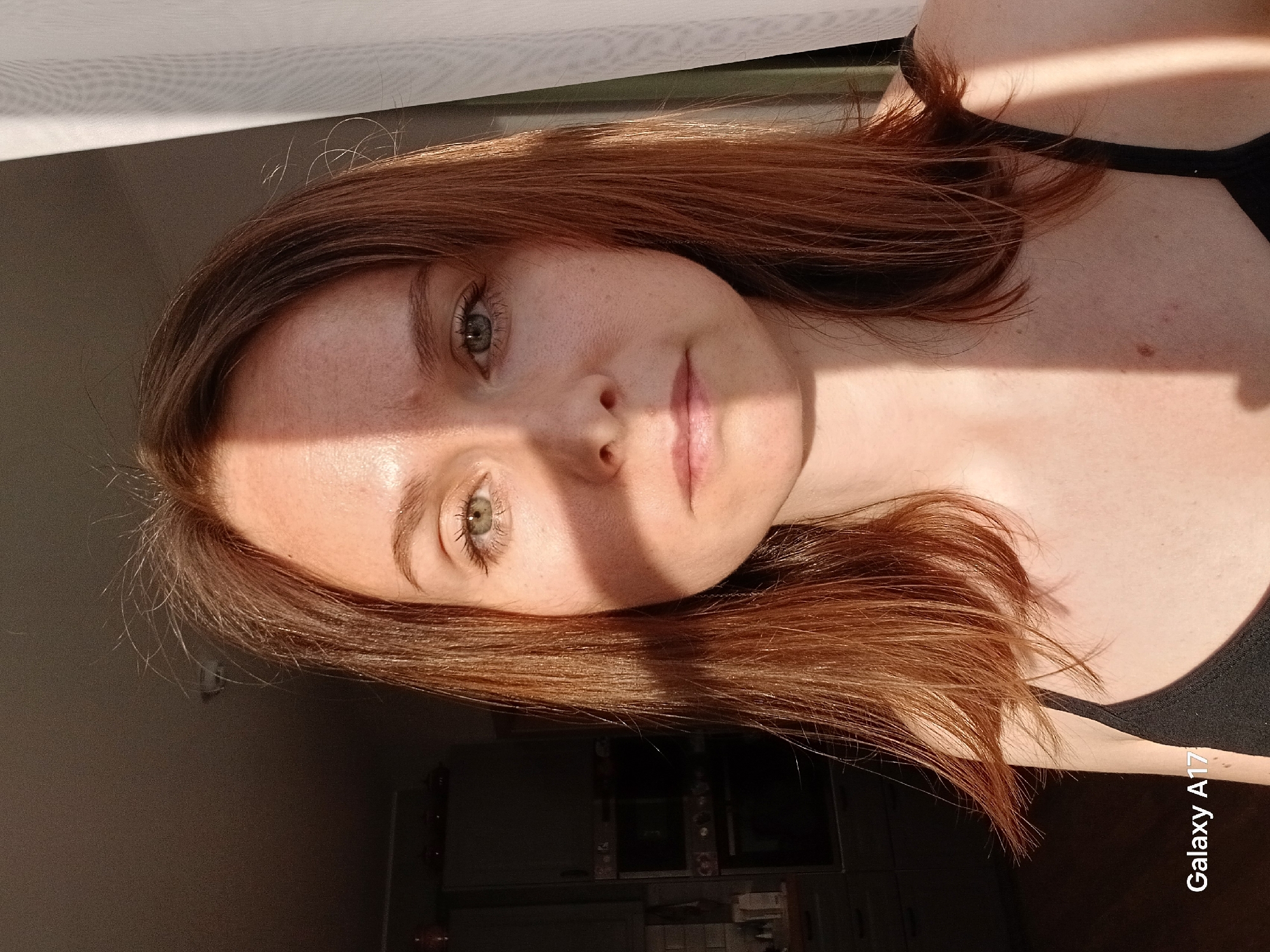 The image shows a fair-skinned person with freckles and auburn hair, facing the camera. Light is casting shadows across their face. They are wearing a black garment. The background is a mix of light and shadow. The location and city in Russia cannot be determined from the visual data.