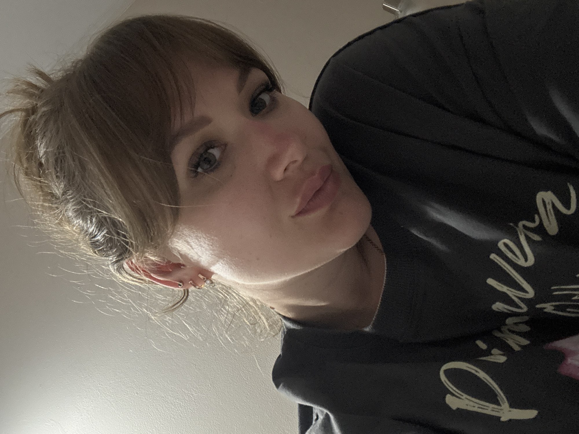 A close-up, low-angle selfie shows a woman with fair skin, light brown hair, and bangs. Her hair is styled in a messy bun. She is wearing a dark grey sweatshirt with white writing. She has multiple piercings in her left ear. The background is a plain, light-colored wall. The location in the city of Yevpatoriya, Ukraine, cannot be determined from the image.