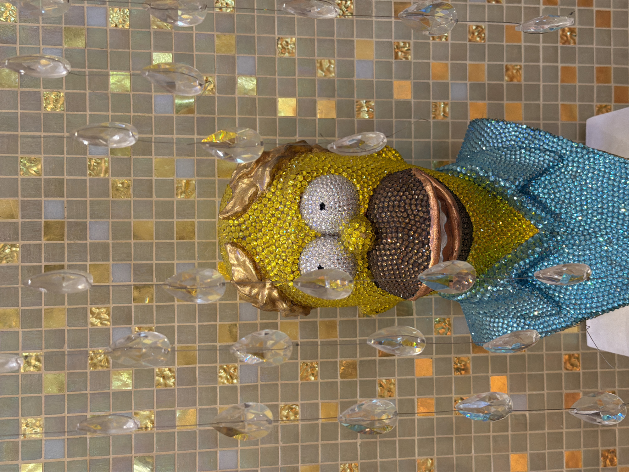 A bedazzled bust of Homer Simpson stands against a backdrop of tan, gray, and gold mosaic tiles. Clear, teardrop-shaped crystals hang on thin strands in front of the bust. The bust's head is yellow, the eyes are white, and the beard is brown, with each element covered in small gems. Homer wears a blue collared shirt. The image might be taken in Moscow, Russia, but there's no definitive evidence of that in the photograph.