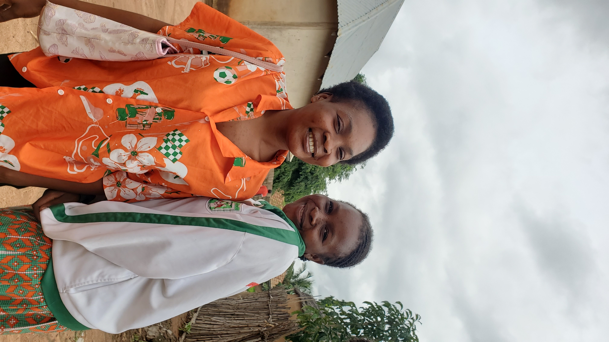 Two young women are posing for a photo outdoors. The woman on the left is wearing an orange button-up shirt with a floral and soccer ball pattern. She has short, dark, curly hair and is smiling broadly. She is also carrying a light-colored purse with a patterned design over her left shoulder. The woman on the right is wearing a white jacket with green trim and a green and orange patterned skirt. She has her dark hair in braids and is smiling. They are standing in front of a simple, light-colored building with a corrugated metal roof. To the right of the women, there is a wooden fence and some greenery. The sky is overcast. The scene suggests a rural or semi-urban environment, possibly in the city of Jalingo, Nigeria.