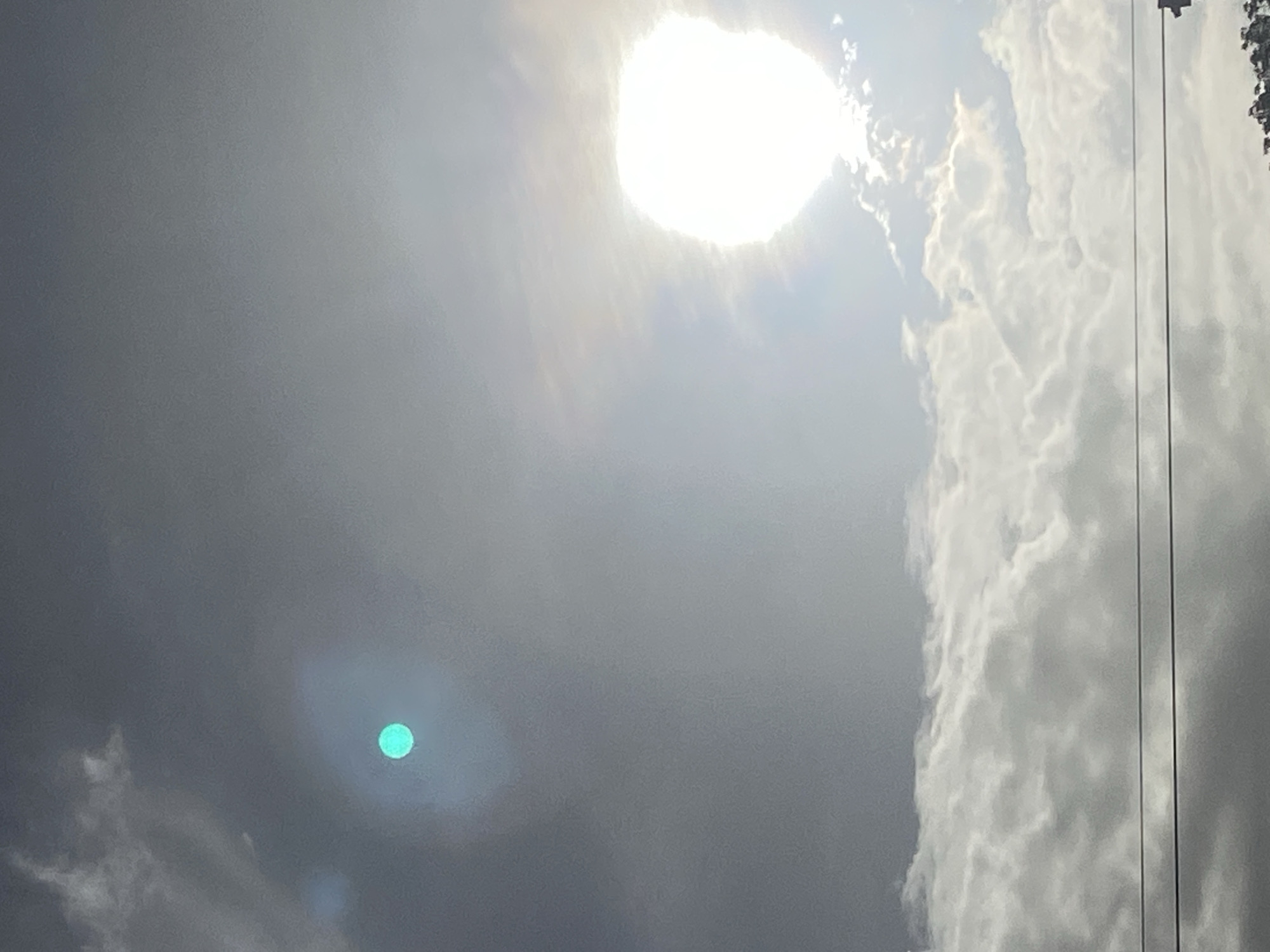This media file captures a bright, daytime view of the sky over Tinaquillo, Venezuela. The sun is the most prominent subject, appearing as an intensely bright, almost overexposed white orb in the upper right portion of the frame, radiating light through a hazy sky.
The sky exhibits varying cloud conditions. On the right side, large, well-defined white clouds are visible, brightly illuminated by the sun, indicating clear patches. However, much of the remaining sky, particularly towards the left and bottom, is covered by a more diffused, grayish haze or thinner cloud layer.
A distinct circular green/teal orb, characteristic of a camera lens flare, is visible in the lower-left quadrant of the image, reflecting the bright sunlight. Along the far right edge of the photograph, several thin, dark vertical lines, resembling power lines or cables, indicate the presence of human infrastructure. At the very top right, there are faint, indistinct dark shapes that could be distant structures or trees. The overall impression is a sunny day with scattered clouds and atmospheric haze. There is no visible text in the image.