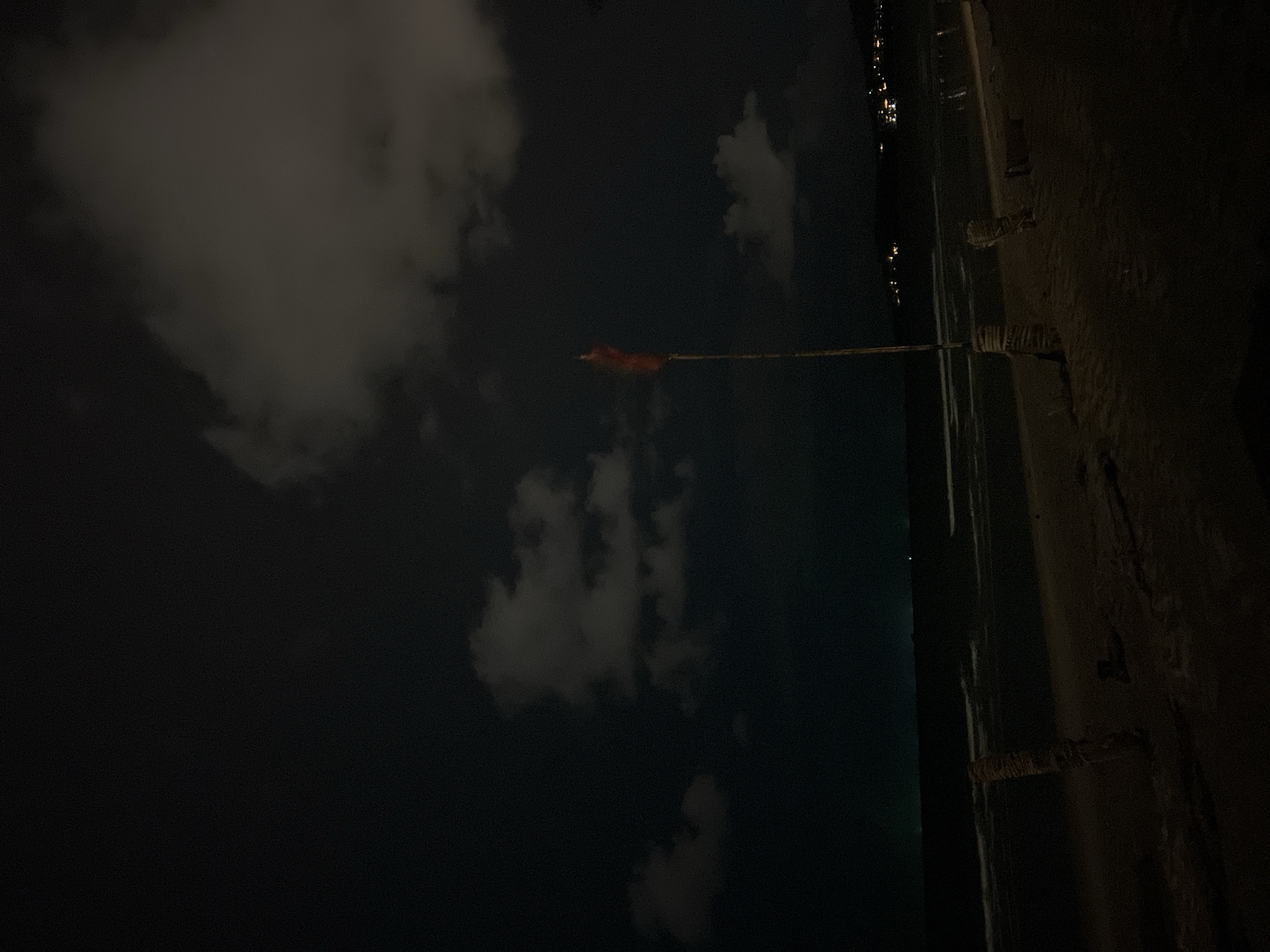At night, a beach scene unfolds with dark, textured clouds overhead and a faintly lit sea in the distance. The foreground features a sandy beach with two weathered posts wrapped in rope. A thin flagpole stands upright, topped with a small, red flag that is slightly blurred, suggesting movement from the wind. In the far distance, a faint line of lights indicates a settlement or town along the coast. The overall atmosphere is dim and serene.