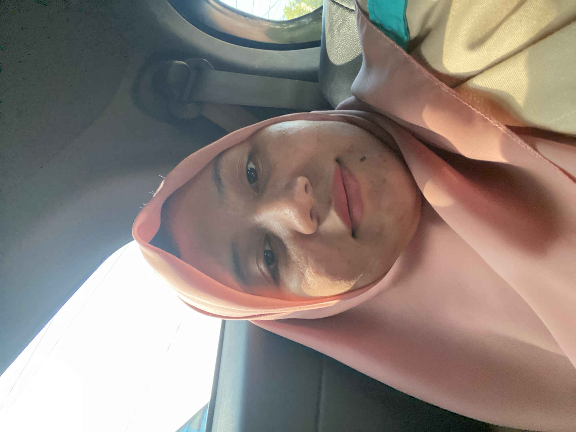 A woman wearing a pink hijab is taking a selfie in the backseat of a car. The car interior is visible, with a seatbelt and the ceiling of the car. The background outside the car windows shows a bright sky and some greenery, suggesting it is daytime. The woman's face is the primary focus, and she appears to be looking at the camera. The scene is consistent with a person traveling in a vehicle within the city of Antasan Senor Ilir, Indonesia.