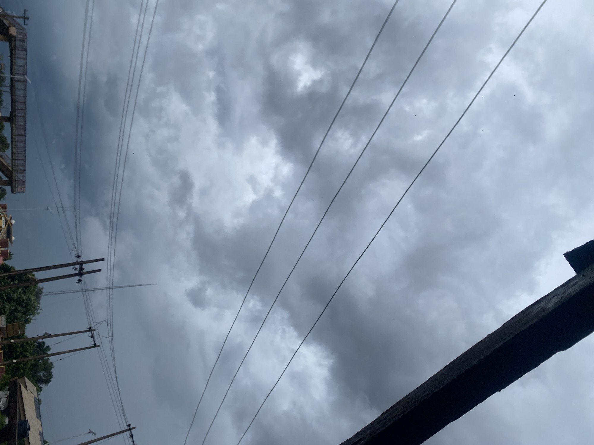 The image displays a heavily overcast sky dominated by dense, dark grey clouds, suggesting stormy or impending rainy weather. Multiple utility lines, likely power or telephone lines, stretch diagonally across the frame from the middle-left towards the upper-right.
On the left side of the image, several wooden utility poles are visible, supporting these overhead lines. Behind the poles, glimpses of green foliage from trees or bushes, and parts of buildings or structures (including what appears to be scaffolding or a building facade), can be seen, indicating an urban or semi-urban environment. A dark, angled structure, possibly a roof edge or a wooden beam, occupies the bottom-right corner of the frame. Small, dark specks, potentially birds or insects, are discernible against the brighter areas of the clouds.
The scene conveys a somber, dim daytime atmosphere under a thick cloud cover. The weather is the prominent feature, hinting at a recent downpour or an impending storm. The visible infrastructure—utility poles and lines—situates the scene in a populated area. This imagery reflects a cloudy day in Auchi, Nigeria. There is no discernible text within the image.