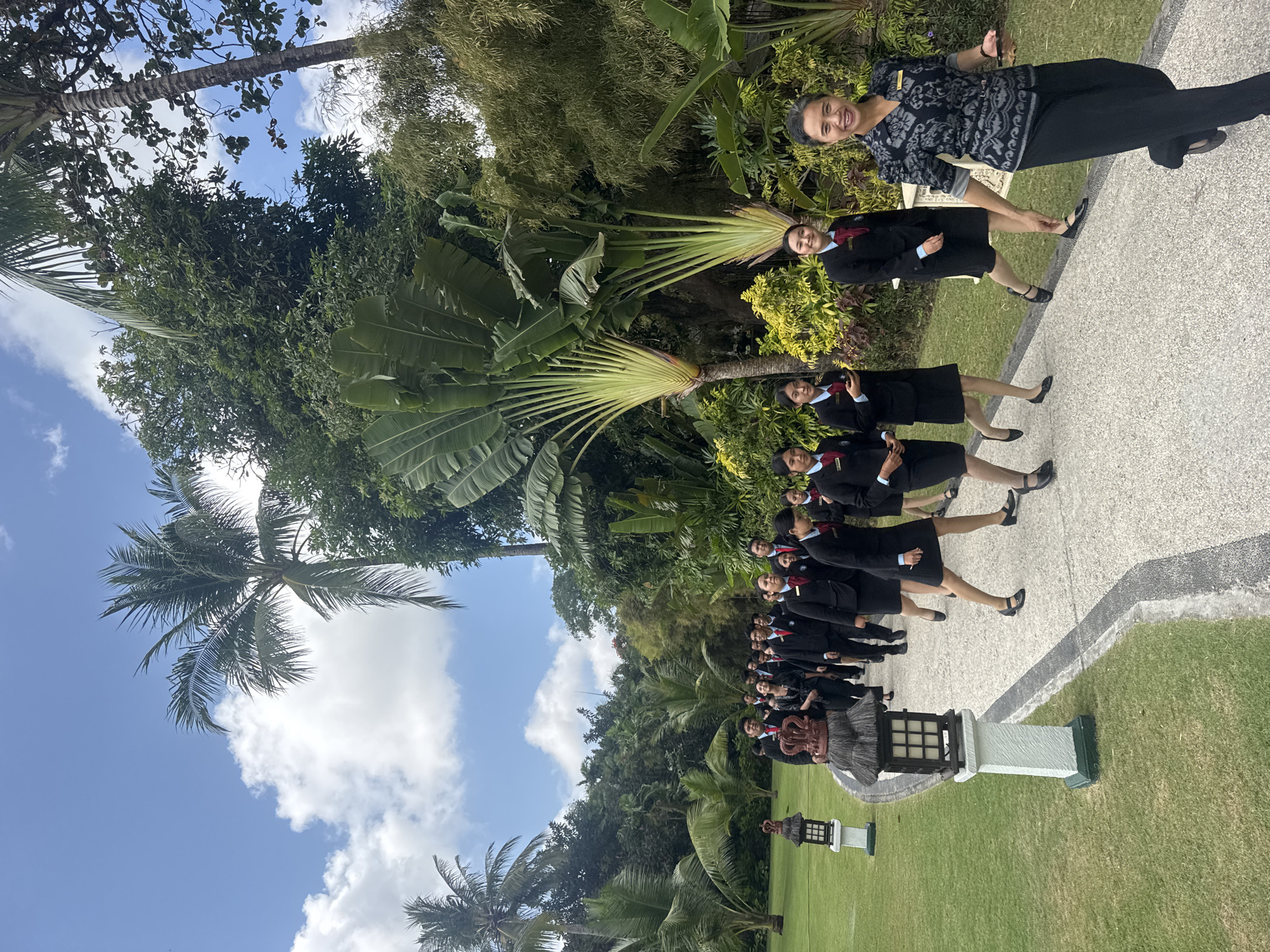 A group of young people, appearing to be students or trainees in hospitality uniforms, walk along a paved path in a lush, tropical environment. The uniforms consist of black blazers, dark skirts or trousers, white shirts, and red neckties or scarves. The group is led by a woman in a patterned black and gray top and black trousers, who is looking back and smiling at the camera. Behind them, a line of palm trees and other dense green foliage dominate the background, suggesting a resort or garden setting. The sky is blue with scattered white clouds. The paved path is edged by a manicured green lawn. Two decorative lanterns, one on a white pillar and another with a thatched roof, are visible on the left side of the path. The scene likely depicts a tour or educational activity at a location in Kuta Selatan, Indonesia, known for its tropical climate and resorts.