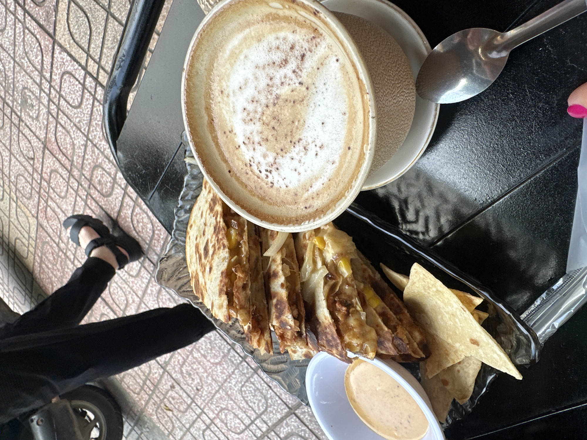 A close-up, overhead shot shows a black tray on a textured black surface, likely a table. On the tray are a stack of sliced quesadillas, a small white bowl of dipping sauce, and a cup of coffee. The quesadillas are golden brown and appear to be filled with cheese and some type of meat. The dipping sauce is a light orange color. The coffee is in a light brown textured mug and is topped with foamy milk and a sprinkle of cinnamon. A silver spoon rests to the right of the coffee cup. In the background, a person's legs, clad in black pants and black sandals, are visible. The ground appears to be paved with square tiles. A portion of a dark-colored wheel, possibly from a motorcycle, is visible on the left side of the image. The overall scene suggests an outdoor seating area, potentially a cafe, in a city like Nha Trang, Vietnam, given the tiled ground and casual outdoor dining setup.