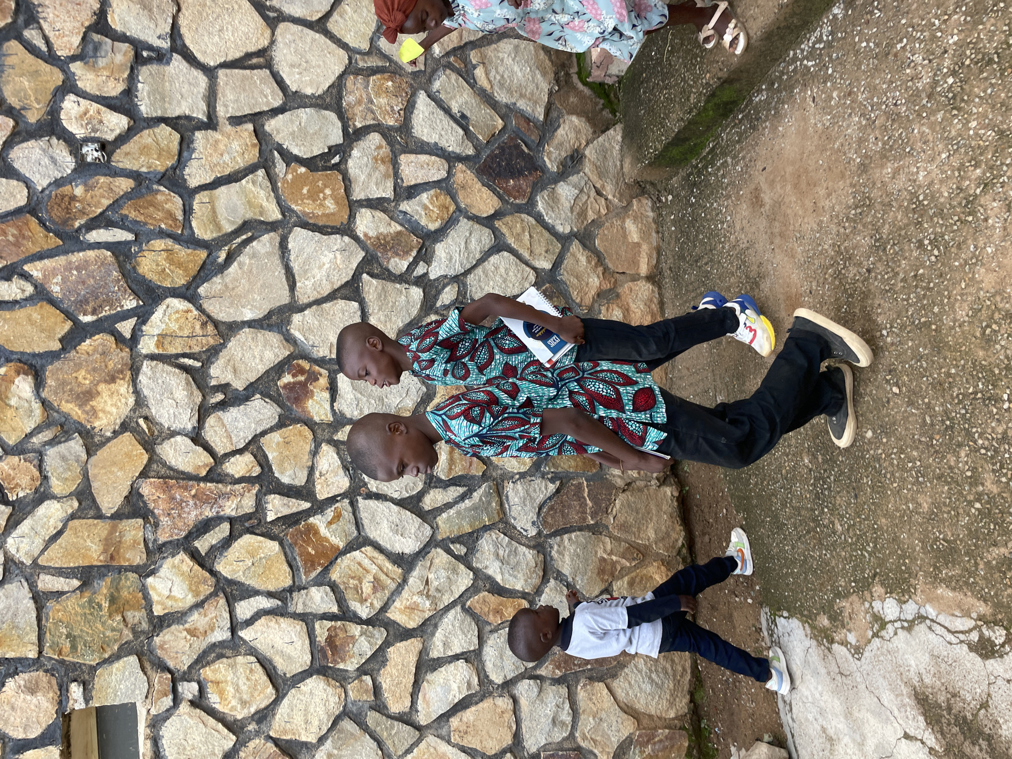 Four children are captured in a photograph taken outdoors, likely on a pathway. The background is dominated by a rustic stone wall constructed from irregularly shaped, natural stones of varying sizes and colors, primarily shades of beige, brown, and grey, interspersed with dark mortar. A small, rectangular window is visible in the upper left portion of the wall.
In the foreground, three boys and one girl are depicted. The boy on the far left, wearing a white long-sleeved shirt over a black long-sleeved shirt and dark trousers, is facing left and pointing with his right hand. He is also wearing white sneakers with light blue and orange accents.
To his right, a second boy is walking towards the left, looking down and to his left. He is dressed in a vibrant, patterned short-sleeved shirt with red, blue, and teal designs, paired with black trousers and black sneakers with cream soles. He is holding a spiral-bound notebook.
A third boy, positioned slightly behind and to the right of the second boy, is also walking to the left. He wears a similar patterned shirt and black trousers, and his footwear consists of white sneakers with prominent blue and yellow accents.
On the far right, a young girl, partially visible, is walking to the right. She is wearing a light blue floral-print dress and a rust-colored headwrap. She has on white sandals.
The ground is a mixture of cracked concrete and gravelly dirt, with some patches of moss on the concrete. A low concrete ledge runs along the right side of the image. The overall impression is that of an outdoor setting in a locale with a distinctive architectural style, potentially a school or residential area. The photograph appears to be taken in the city of Jos, Nigeria, a region known for its stone architecture and diverse population, which includes a significant Christian and Muslim population, with both communities contributing to the cultural fabric of the city. The children's attire and the naturalistic setting are consistent with a scene in this part of Nigeria.