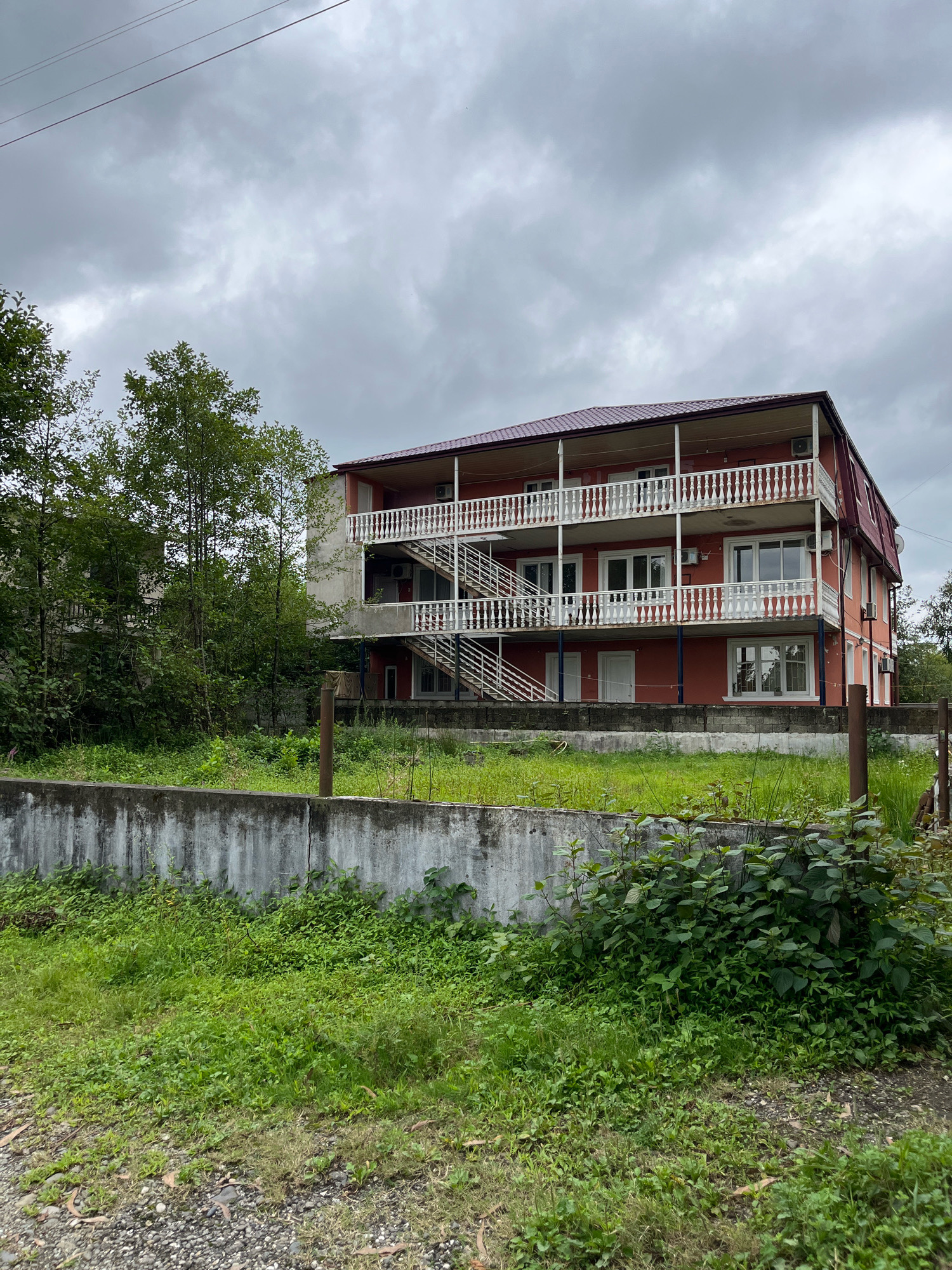 The image shows a multi-story building with a red roof and peach-colored walls, featuring multiple balconies with white railings and an external staircase. The building is situated in Ureki, Georgia, a seaside resort town known for its magnetic sand. The sky is overcast with gray clouds. In the foreground, there is a low concrete wall with lush green grass and vegetation growing around it. The scene appears to be a residential or possibly a small hotel property.