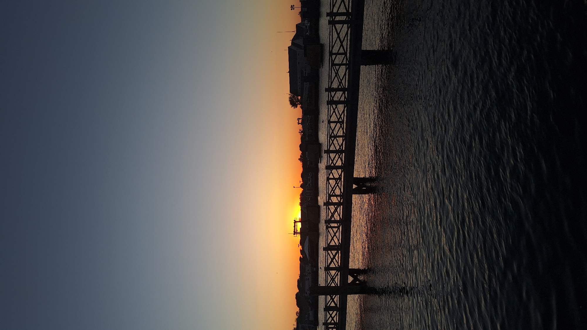 The image depicts a sunset over the water in Nantucket, United States of America. A wooden pier with a crisscross railing stretches across the frame from left to right, supported by dark pilings. The water below the pier is dark and slightly rippled, reflecting the muted colors of the sunset. On the horizon, silhouetted buildings and structures are visible, with the sun setting just above the horizon, casting an orange glow. The sky transitions from a pale orange near the horizon to a deep blue at the top of the frame. There are no people or animals clearly visible in the image. The overall scene conveys a peaceful and serene atmosphere.
