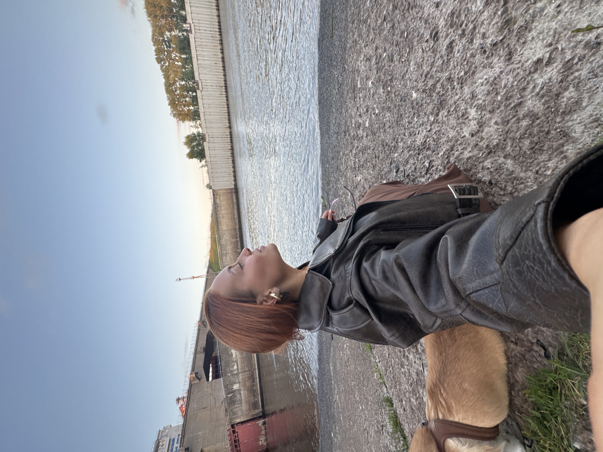 An outdoor, eye-level, selfie shot shows a young woman with short, auburn hair sitting by a body of water. She is wearing a dark brown leather jacket and a brown skirt. Her head is tilted back, and her eyes are closed as she faces the sky. To her left, a small dog wearing a harness is visible. In the background, a concrete structure with red gates stands on the left, and a long, white-paneled wall runs along the right bank of the water. The sky is a pale blue with a few wispy clouds. The setting appears to be a dam or lock system along a river.