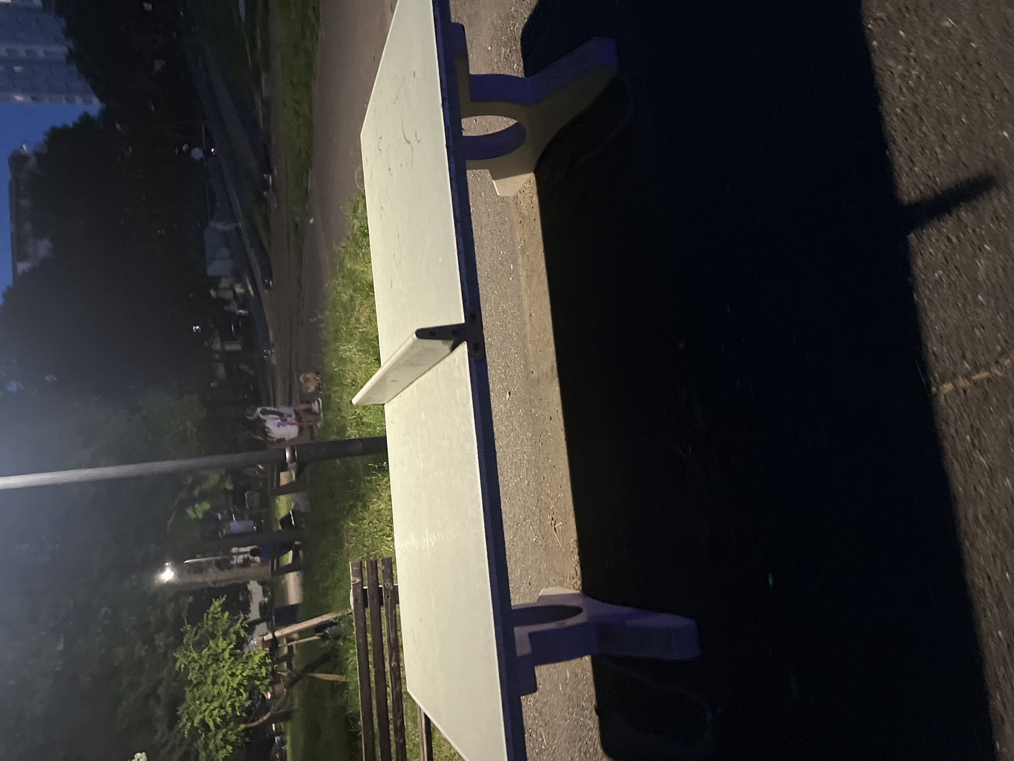The image shows a picnic table in a park. The table is light-colored and has a built-in bench on each side. The table is located on an asphalt path, and there is grass and trees in the background. Other picnic tables are visible in the background, with people sitting in chairs. The photograph may have been taken at night. The location could be Burgas, Bulgaria, but there is no definitive information in the image to confirm this.