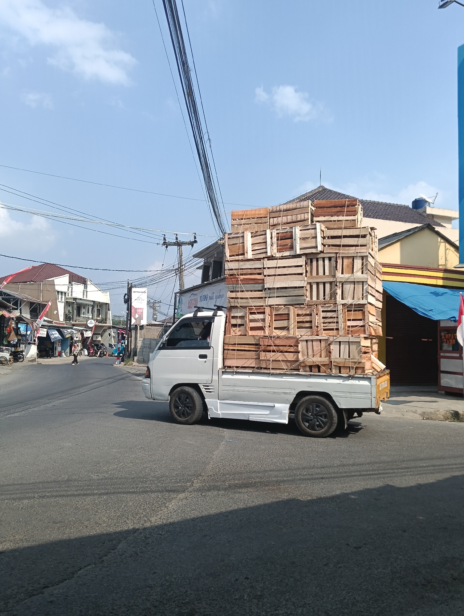 In the city of Puspahiang, Indonesia, a white utility truck is captured in a side-view medium shot, driving on an asphalt road under a clear blue sky with scattered clouds. The truck is heavily laden with a large stack of wooden crates, bound together with ropes. The crates are arranged in a dense, towering structure, filling the entire bed of the truck and extending upwards. The truck's wheels are black. To the left of the truck, buildings line the street, some with awnings and signs, suggesting a commercial area. A few people are visible walking on the sidewalk and crossing the street in the distance. Power lines crisscross the sky above the scene. To the right of the truck, a building with a yellow and red striped facade is visible, partially obscured by a blue tarp. The overall scene depicts a busy street with a significant load of goods being transported.