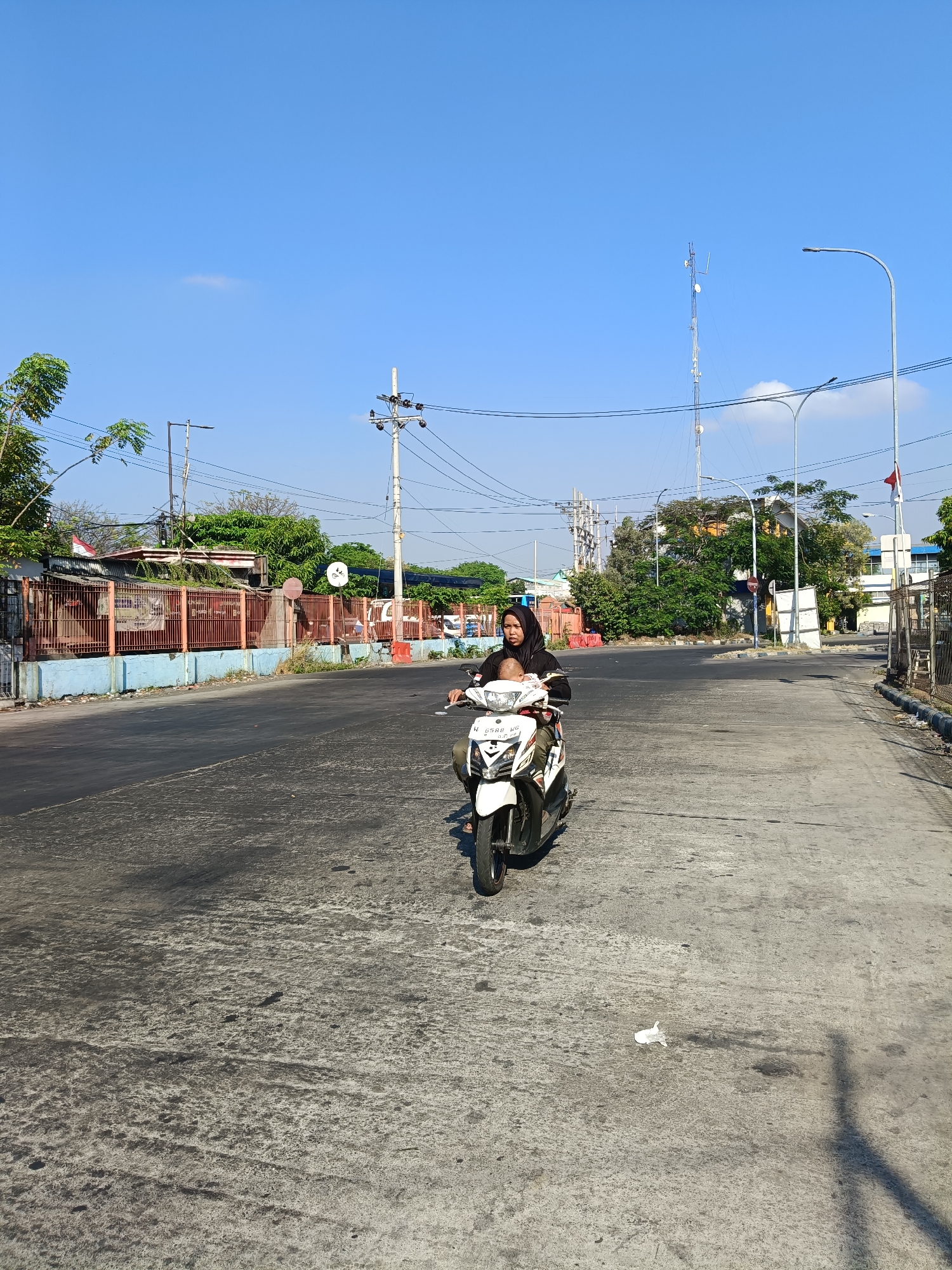 In Kedungrejo, Indonesia, on a clear, sunny day, a woman wearing a black hijab rides a white scooter down a paved road. She is carrying a baby on the front of the scooter. The road is lined with a low blue wall and a metal fence. Behind the fence, buildings and trees are visible. Utility poles and power lines crisscross the sky, which is a bright, cloudless blue. A distant cell tower stands tall against the horizon. The road surface shows signs of wear and tear, with patches of darker asphalt and lighter concrete. A piece of white trash lies on the road in the foreground.
