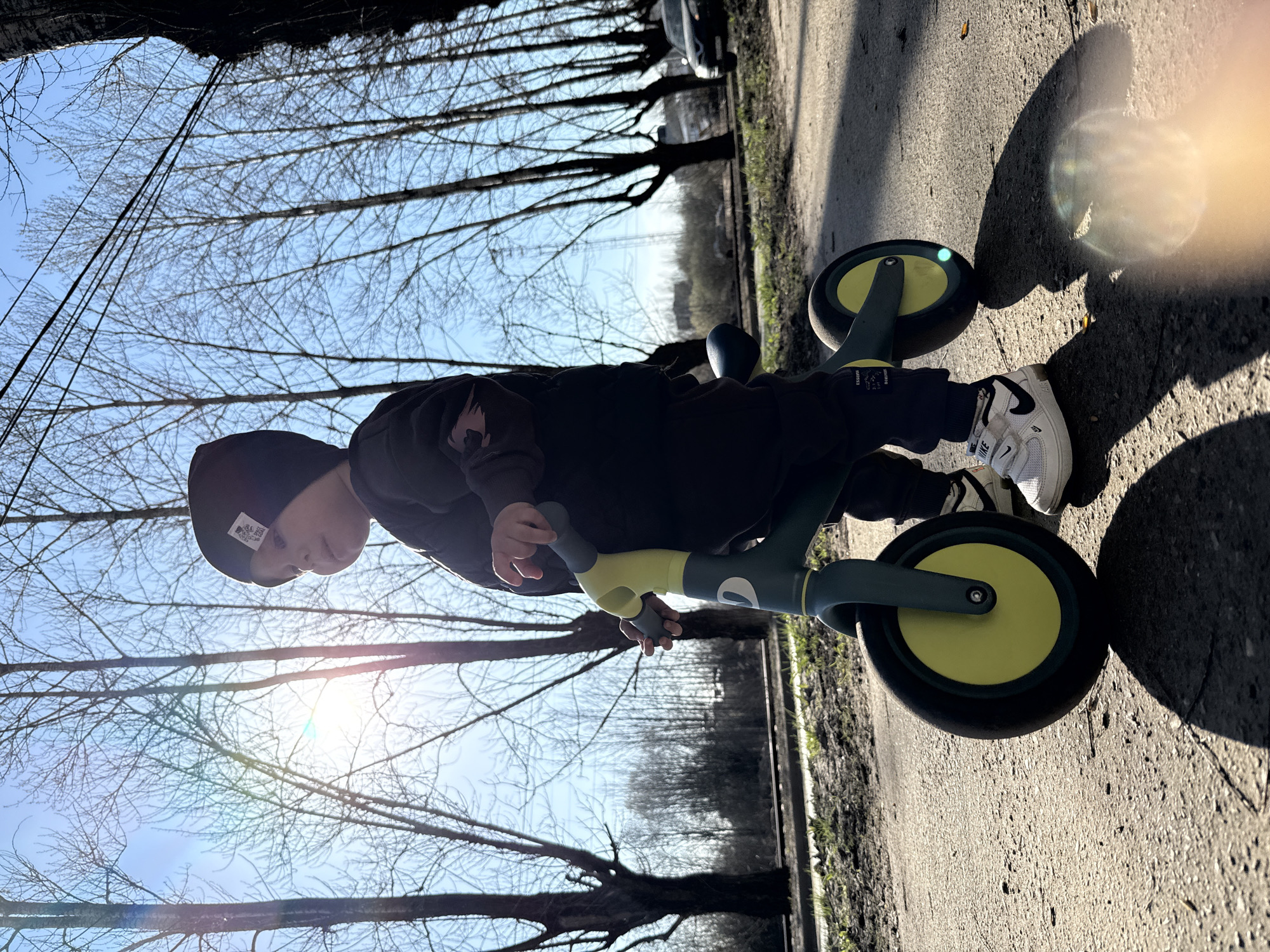 A young child, wearing a black beanie and a dark sweatshirt, is riding a light green and dark grey balance bike on a paved surface. The child wears white Nike sneakers with velcro straps. The child's shadow is visible on the pavement. The pavement has a narrow border of green vegetation on the right. Bare tree branches fill the background and upper parts of the image. A portion of a dark-colored vehicle is visible beyond the trees. The location is Rusanovo, Russia, as implied by the user.